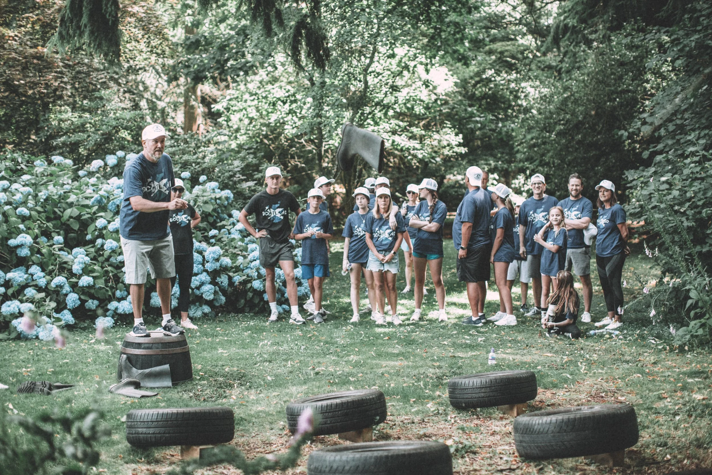 Group of children and adults gathered outdoors in a park-like setting, listening to a man standing on a wooden barrel, with tires placed on the ground in front.