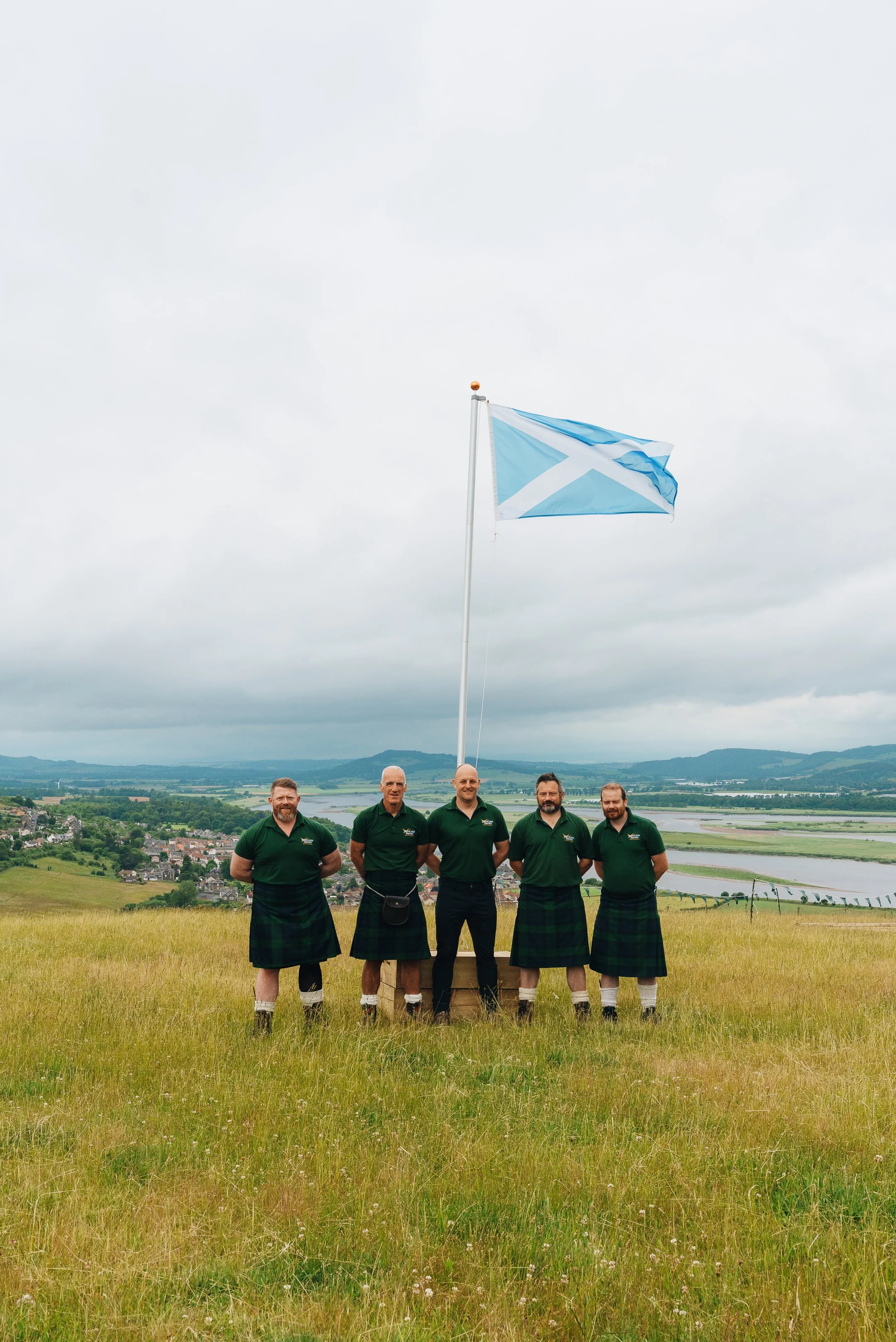 Five men dressed in traditional Scottish kilts and green polo shirts standing in a grassy field, holding a flagpole with the Scottish flag against a scenic landscape with hills, water, and a cloudy sky.