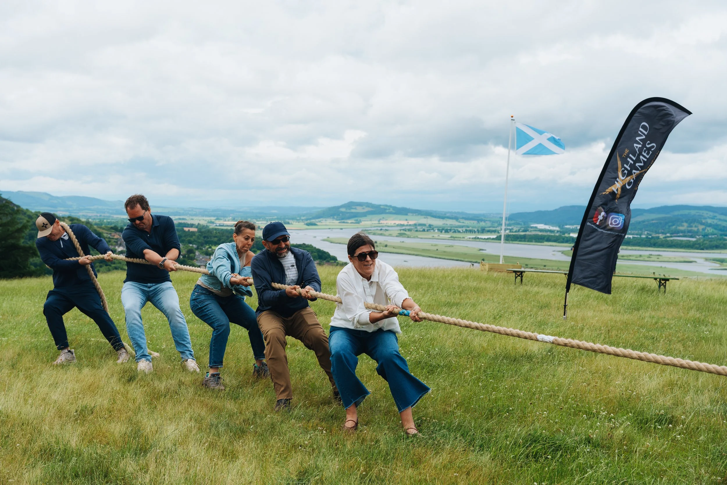 A group of people participating in a tug-of-war game outdoors on a grassy field with hills and a river in the background. There are flags and banners nearby, one indicating support for Highland Games.