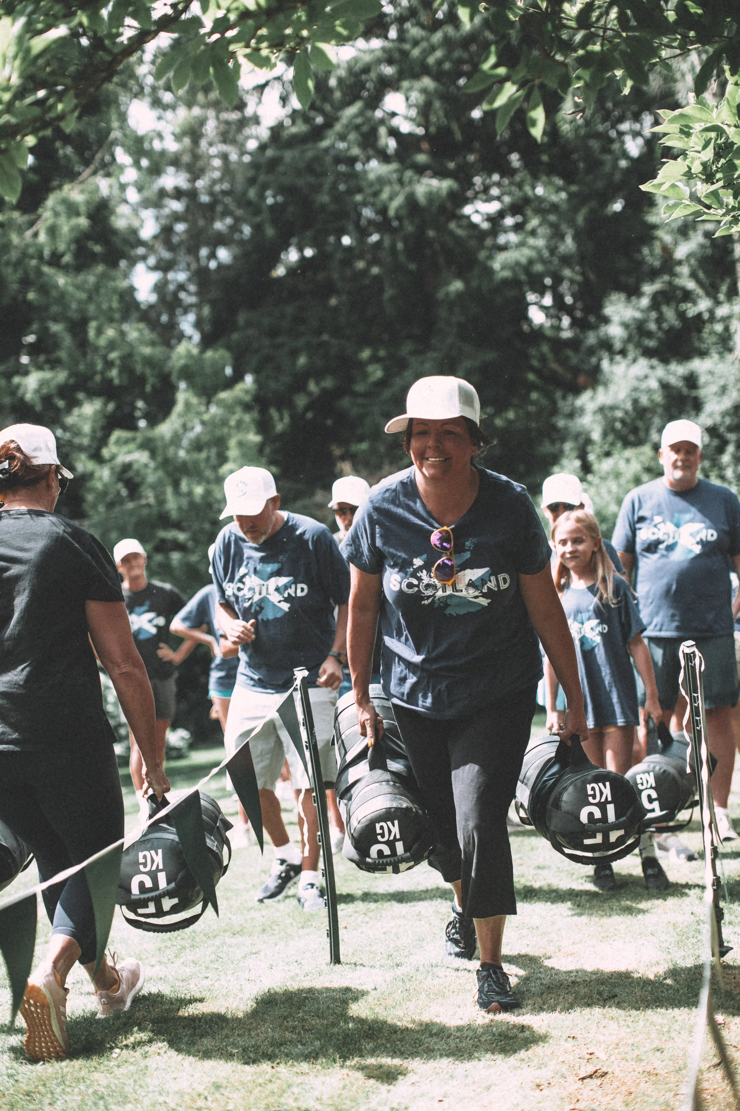 Group of people outdoors, carrying sports bags and gear, walking on grass under trees, wearing matching T-shirts with the word 'Scotland' and a mountain logo, some wearing white caps.