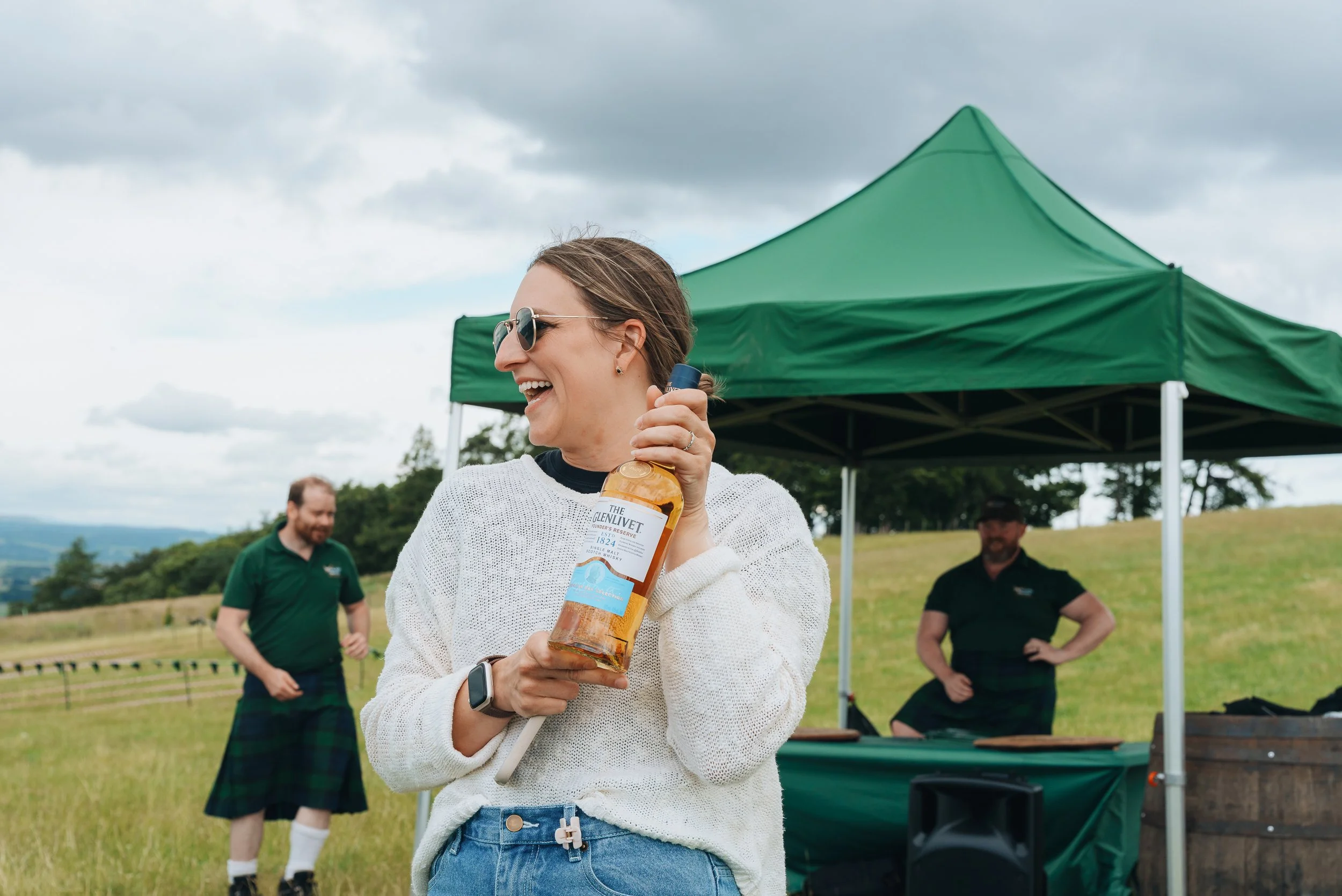 A woman wearing sunglasses and a white sweater holding a bottle of whiskey outdoors at a gathering or event, with two men in kilts and another person in the background near a green canopy tent.