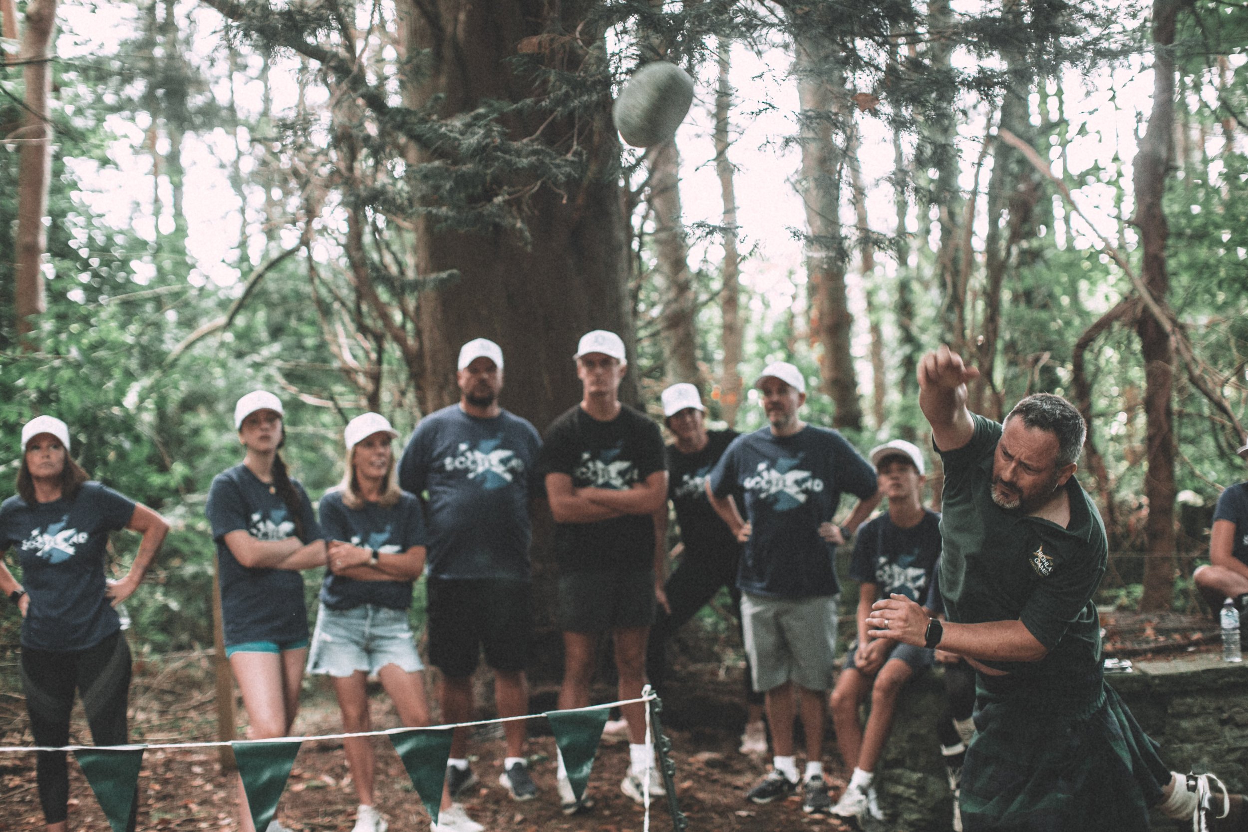 A group of people in a forest, wearing matching shirts and white caps, watching a man throw a rock into the water.