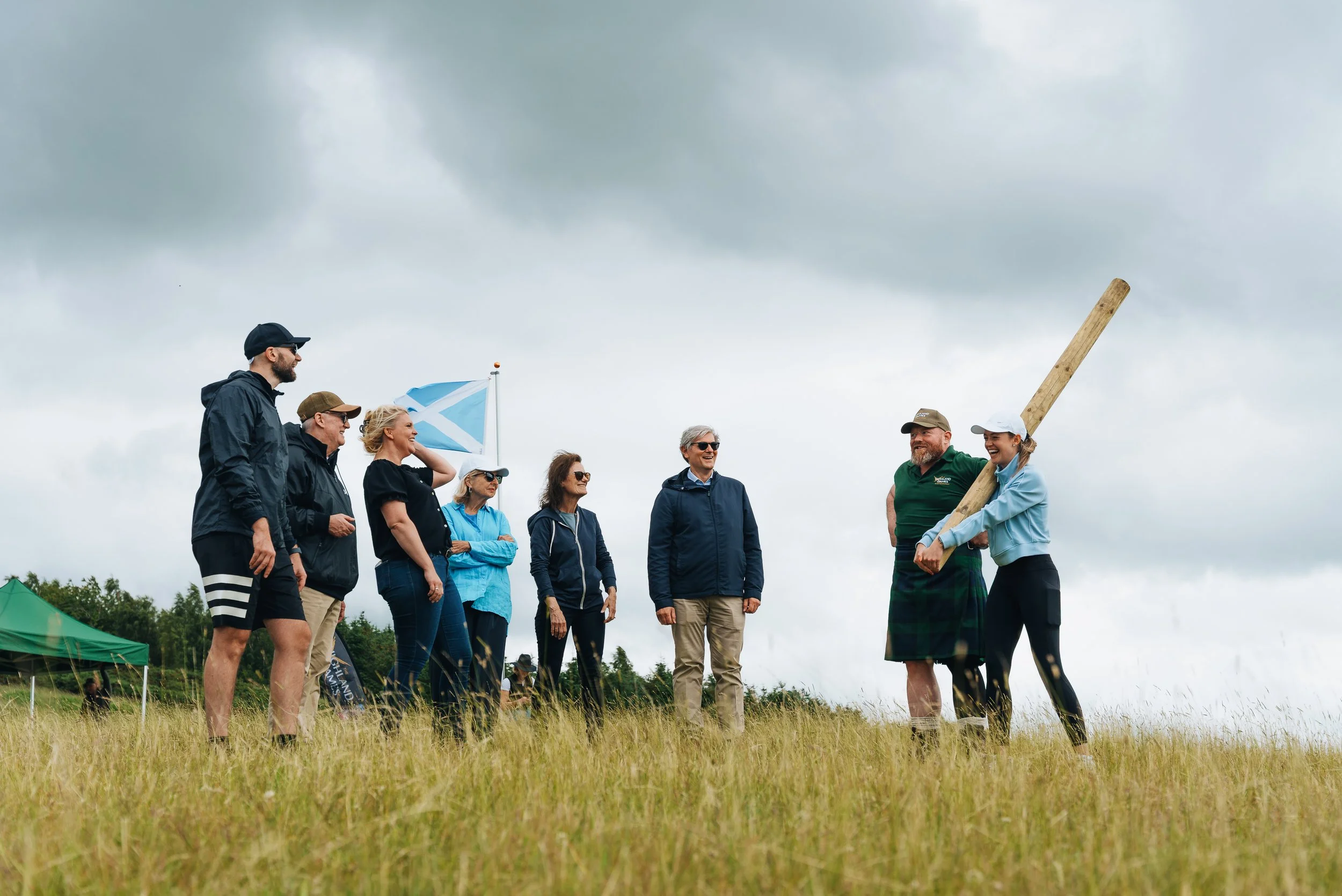 A group of people standing outdoors on a grassy field under cloudy sky, some wearing jackets and sunglasses, while one person in traditional Scottish attire holds a wooden club.