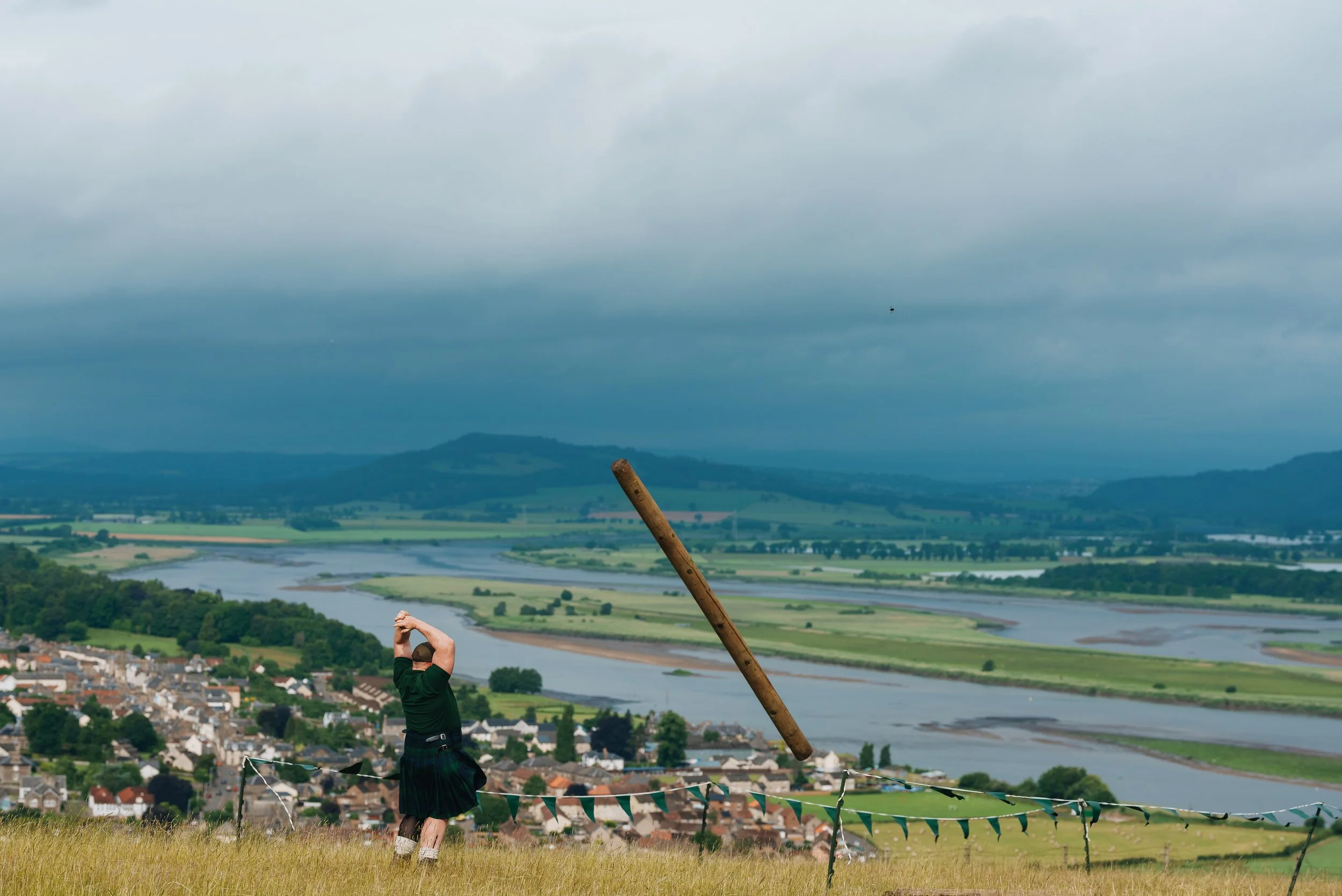 A person in a kilt and green shirt is standing outdoors on a grassy hill, holding a large wooden pole in a throwing stance. The background features a river, fields, and a small town under a cloudy sky.