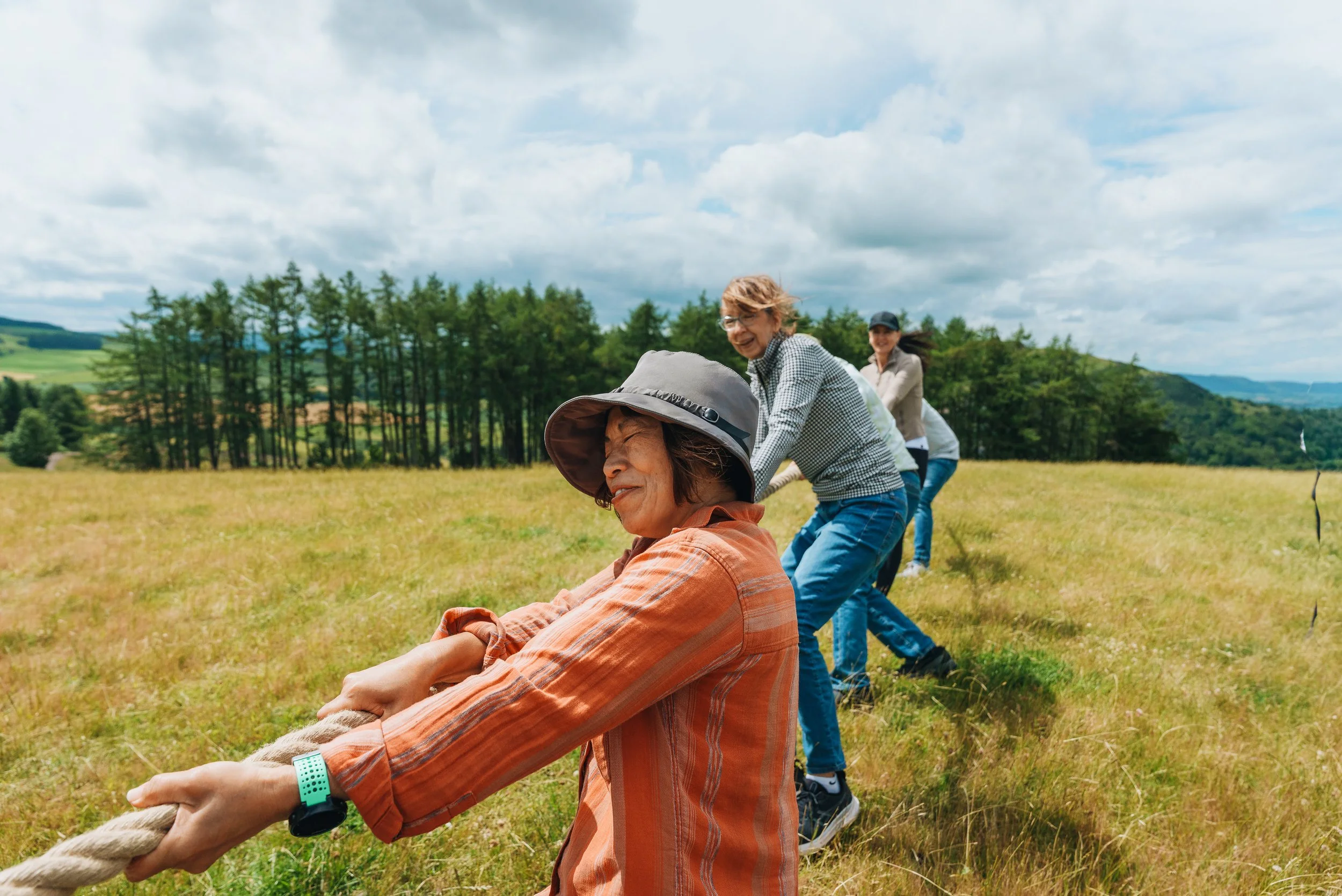 Four women playing tug-of-war outdoors in a grassy field with trees and cloudy sky in the background.
