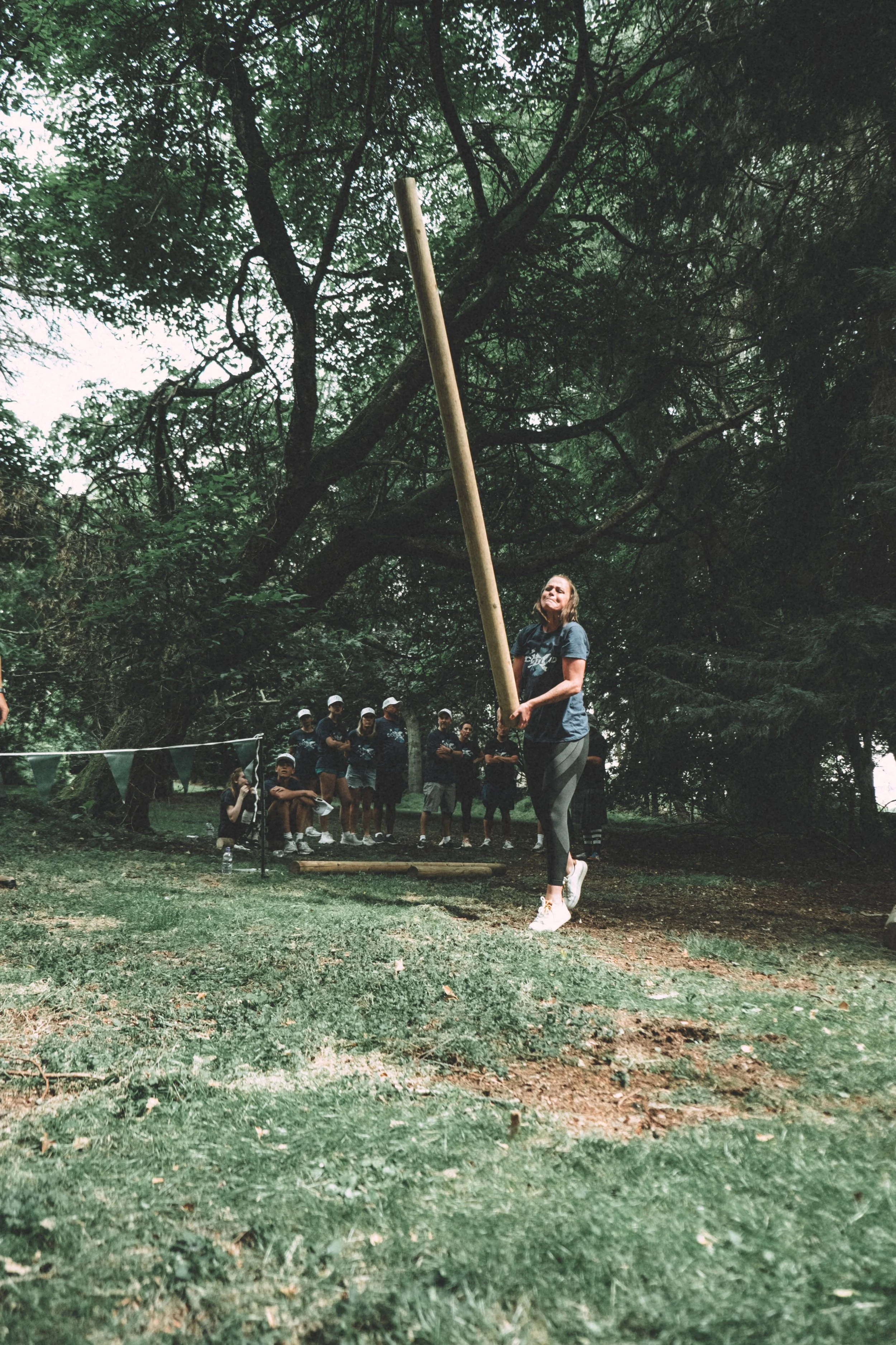 A woman holding a pole stands in front of a group of children and adults outdoors beneath large trees. The group appears to be participating in an outdoor activity or game.