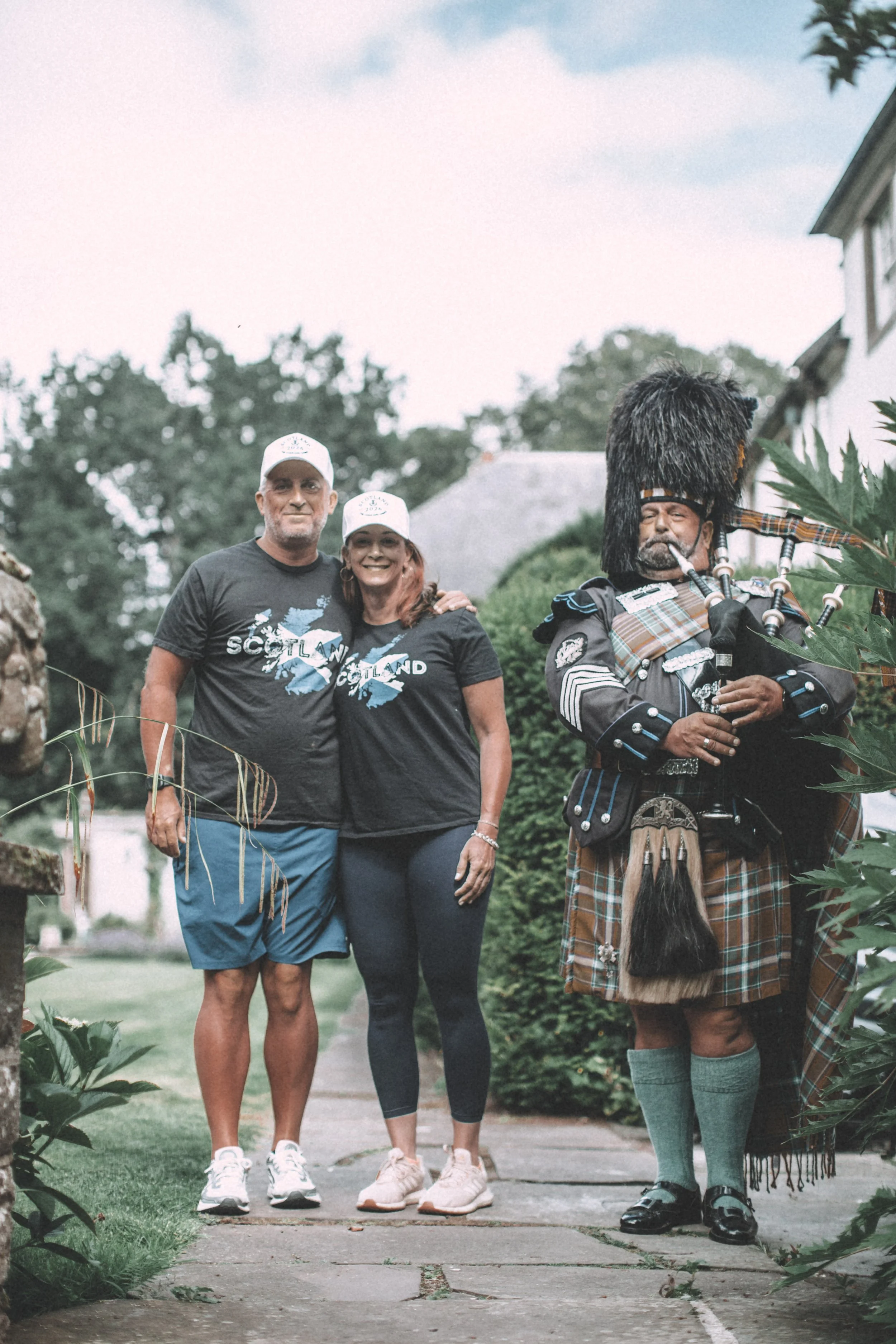Two tourists and a bagpiper standing on a sidewalk outdoors.