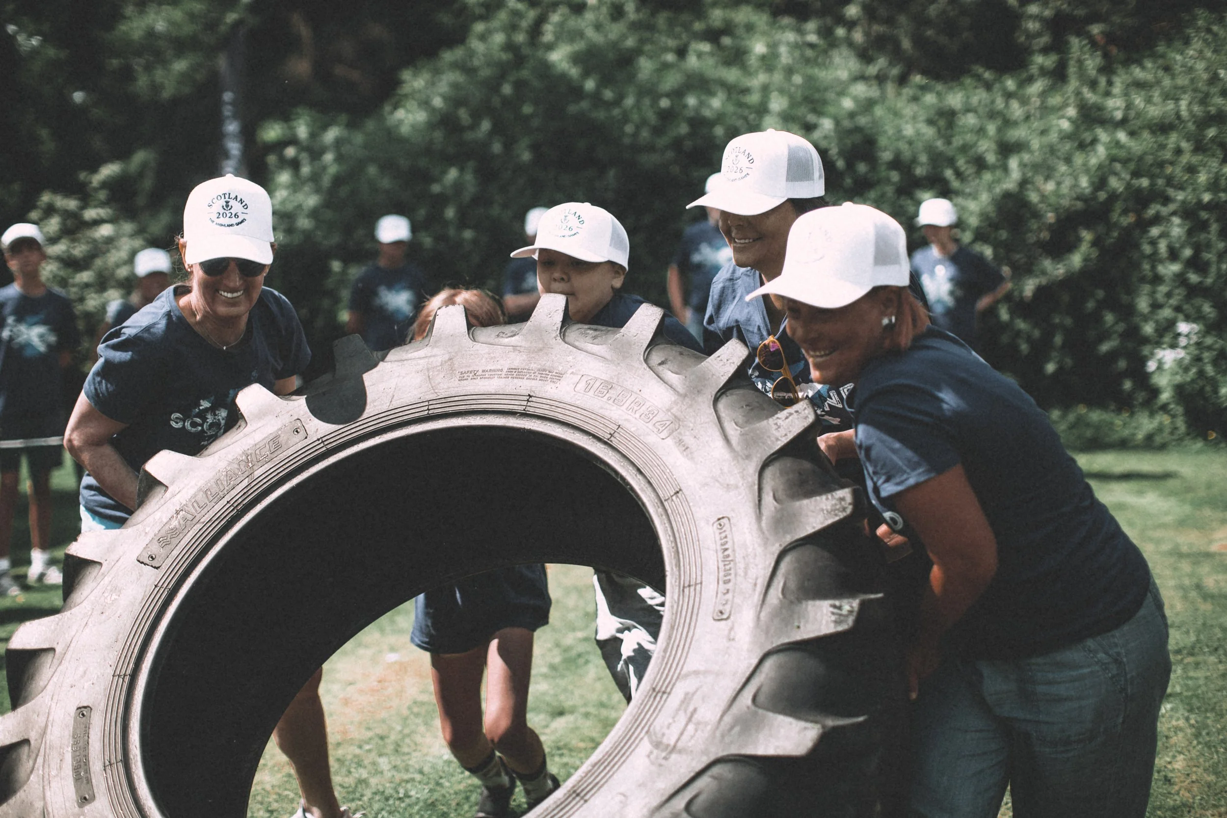 Group of people participating in an outdoor event involving a large tractor tire, wearing white caps and navy shirts, with trees in the background.