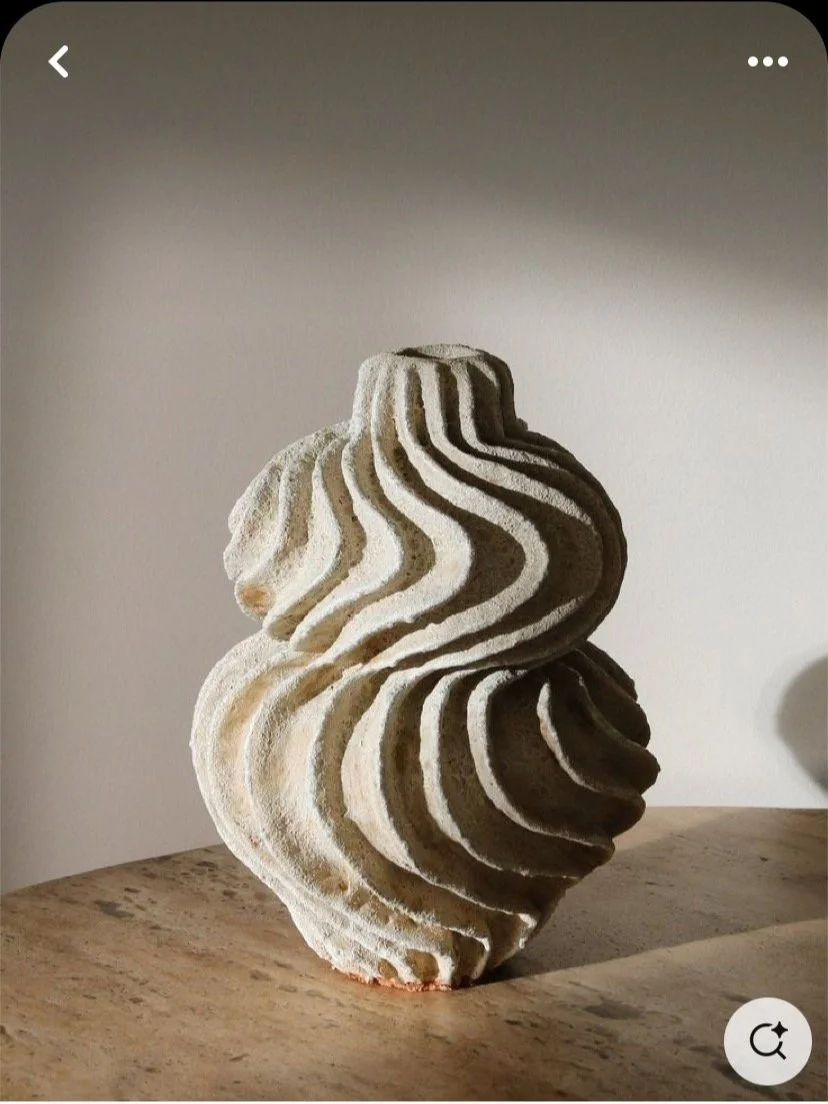 A sculptural piece resembling a stylized human face with flowing, wavy lines, made from a textured material, placed on a wooden surface against a plain wall.