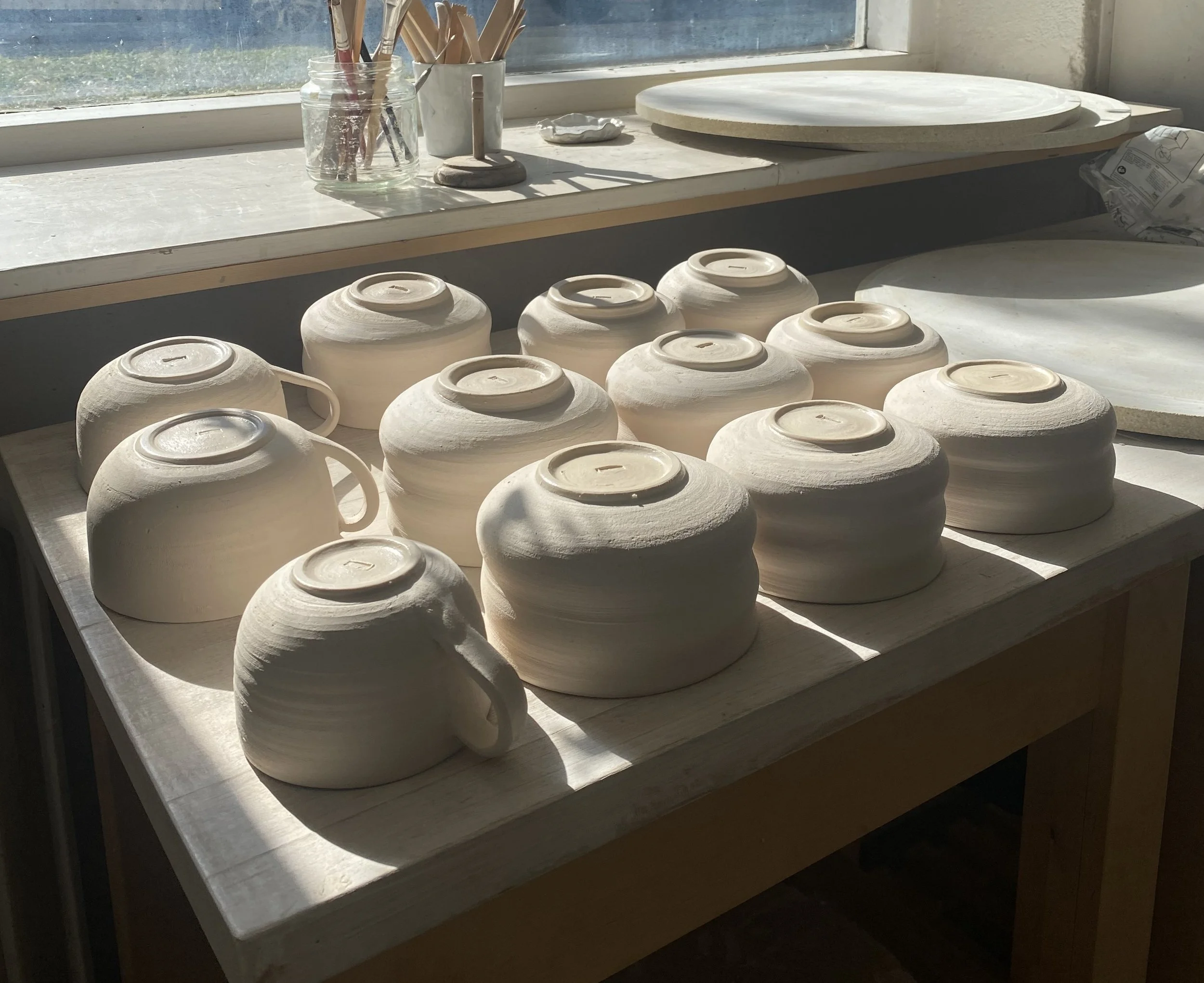 Clay pots upside down on a wooden table in a pottery studio, with a window and art supplies in the background.