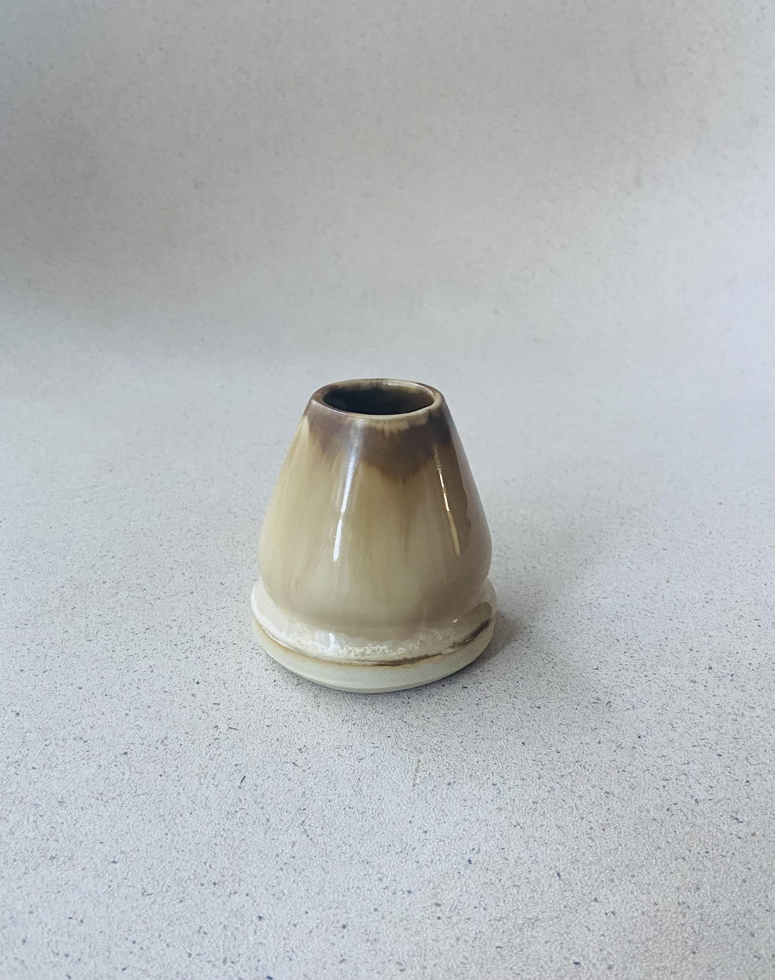 A small, cream-colored ceramic vase with a rounded body and a narrow opening, placed on a speckled gray surface with a plain white background.