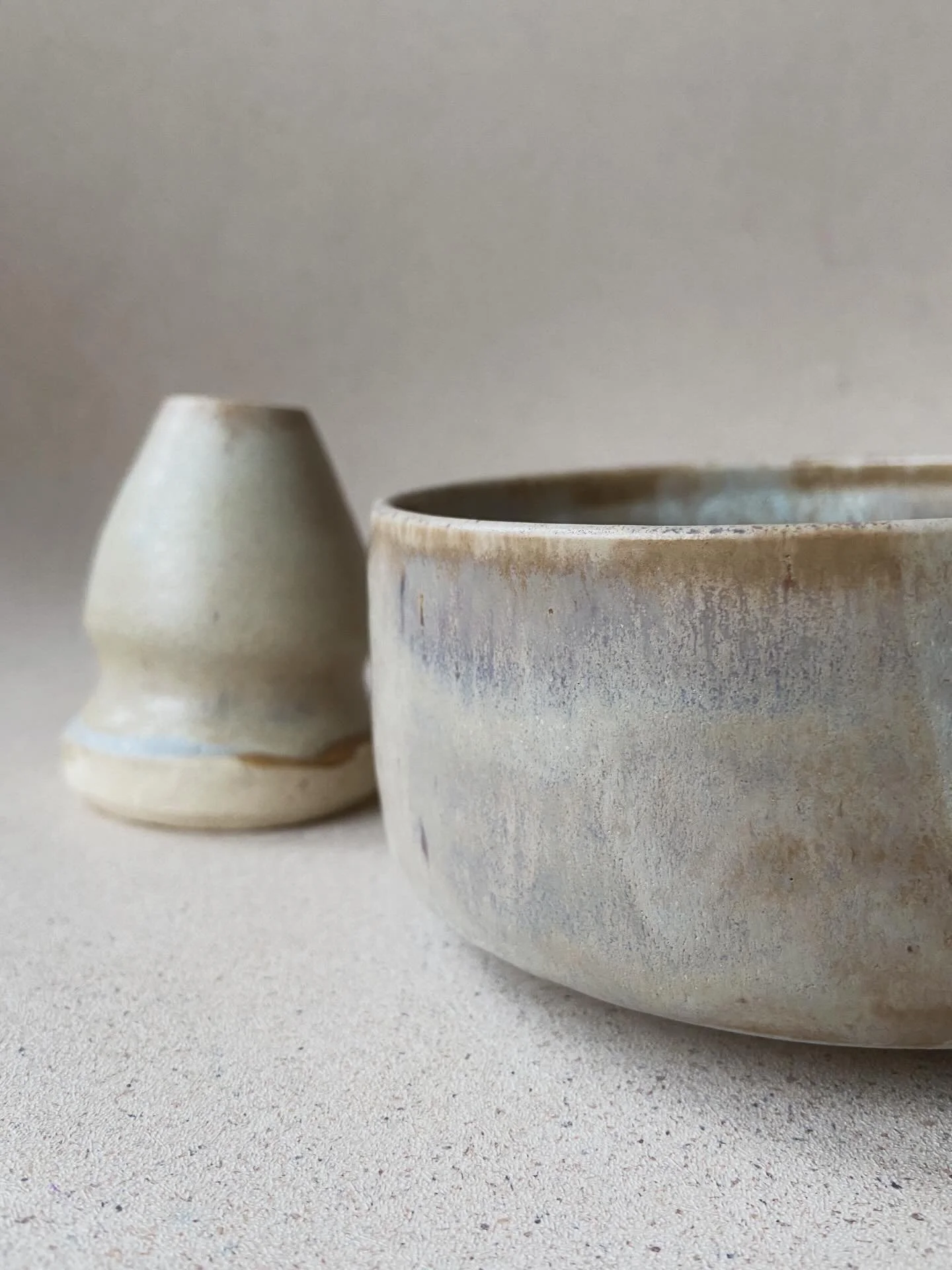 Close-up of two handmade ceramic pottery pieces, a small vase and a bowl, in neutral tones placed on a flat surface with a plain background.