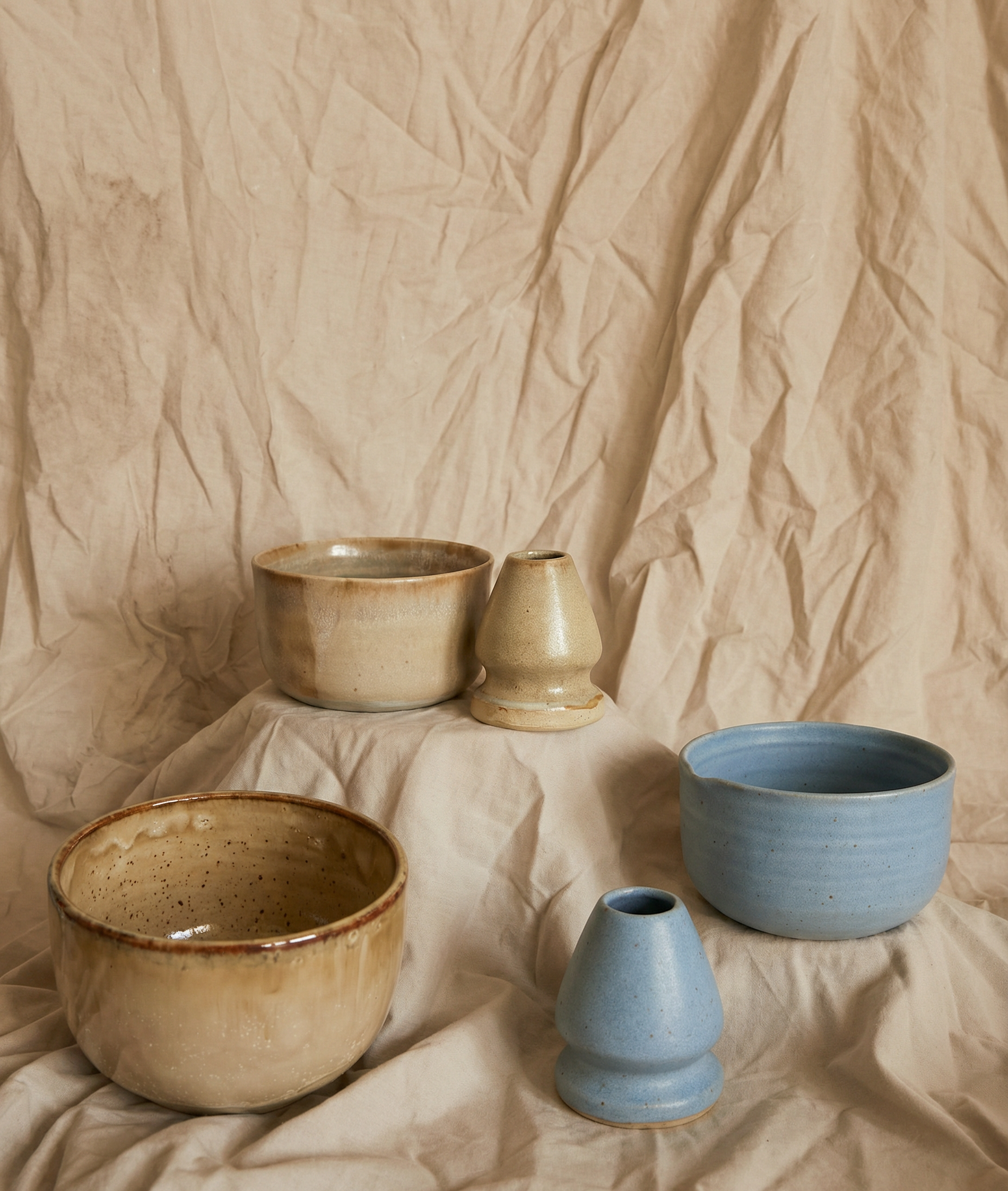 A collection of ceramic bowls and cups in various sizes and colors, set against a crumpled beige paper background.