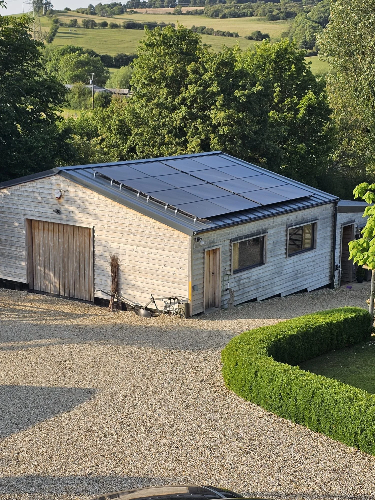 A wooden house with solar panels on the roof, a gravel driveway, a neatly trimmed hedge, and green trees and hills in the background.