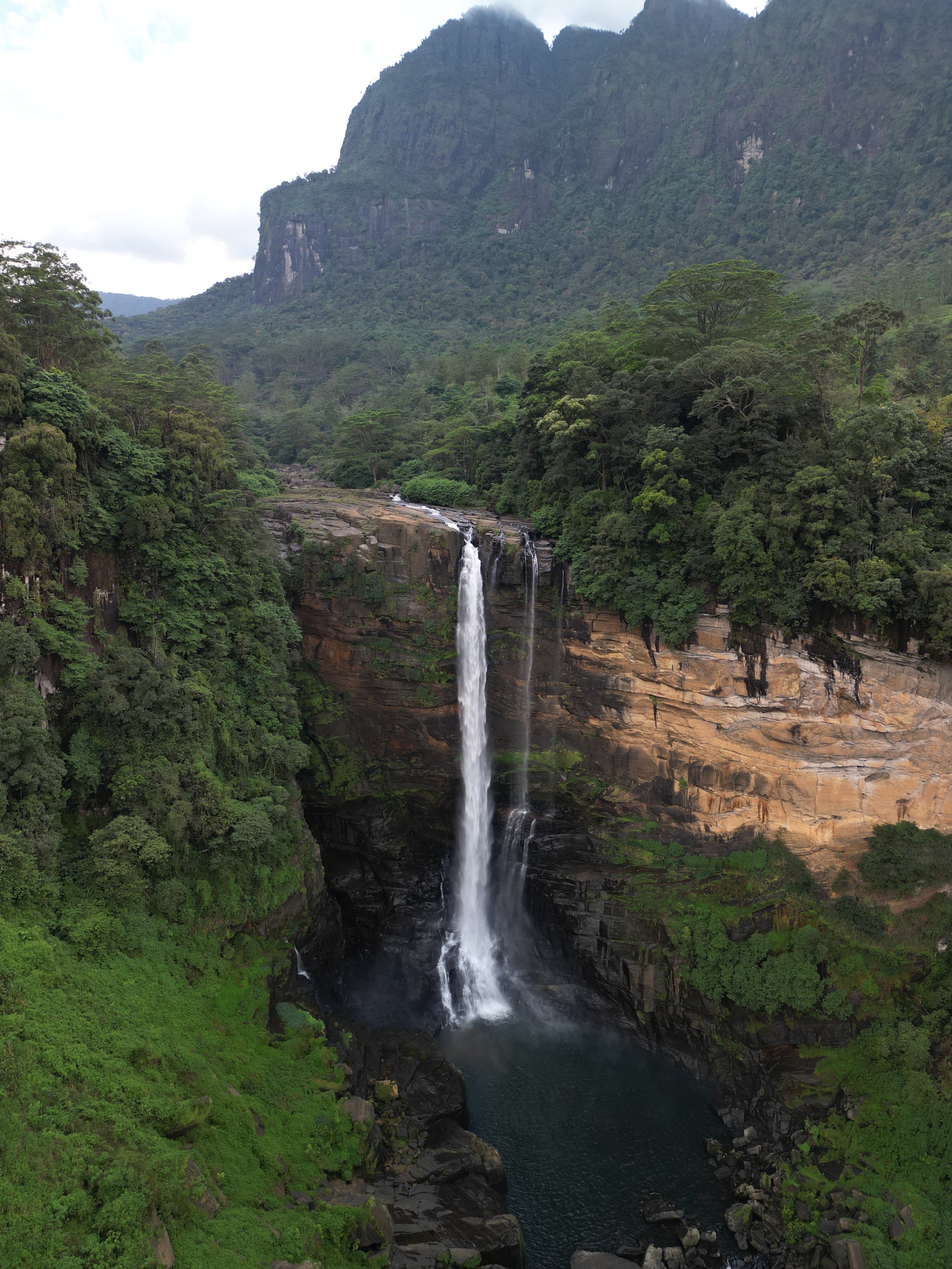 A tall waterfall cascading down a rocky cliff into a dark pool surrounded by lush green forest and mountain in the background.
