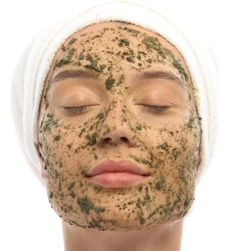 A woman with her eyes closed, wearing a white headband, has a green facial mask applied evenly across her face.