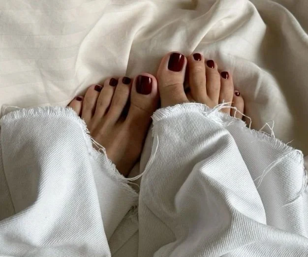 Close-up of a person's feet with red painted toenails, partially covered by white frayed jeans, resting on a white bedsheet.