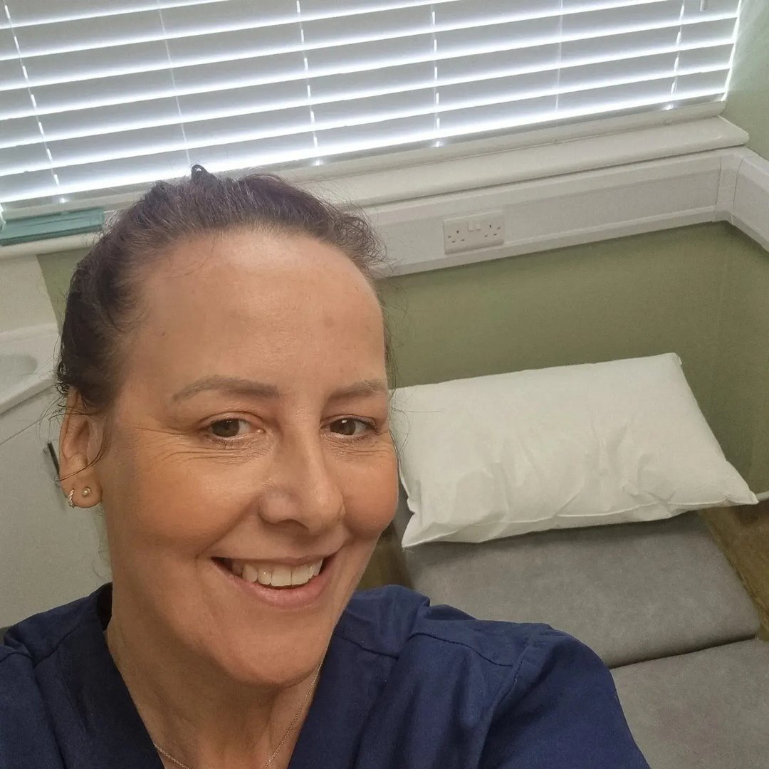 A woman smiling in a medical examination room, with a pillow on a bed, a window with blinds, and medical equipment in the background.