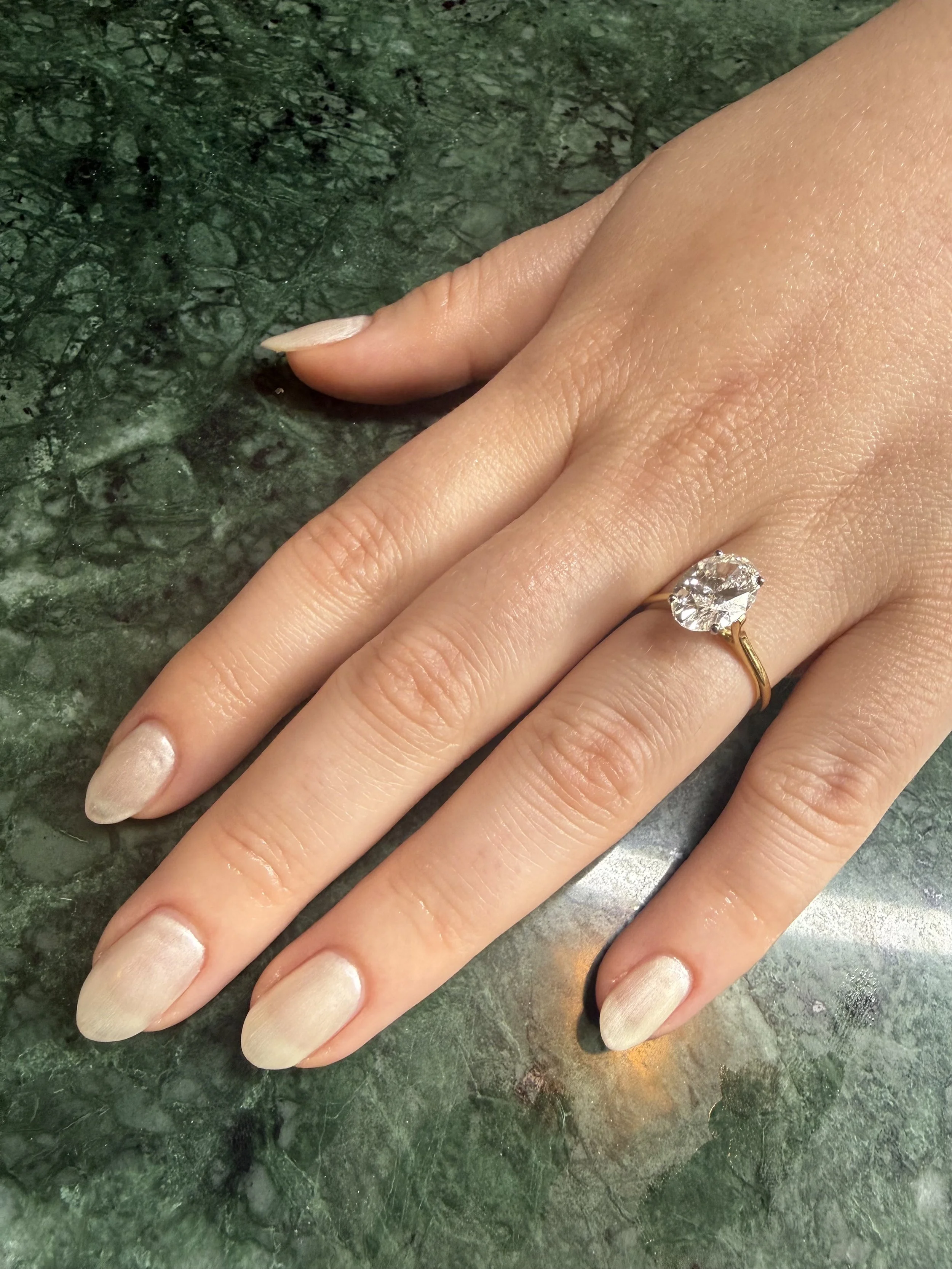 Close-up of a hand after a gel service with manicured fingernails, wearing a large diamond engagement ring, resting on a green marble surface.