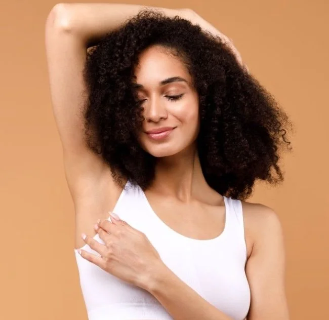 A woman with curly hair posing with her eyes closed, lifting her arm to reveal her underarm against a beige background.