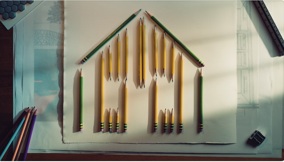 Colored pencils arranged on a sheet of paper to form a house shape, with other drawing tools around it.