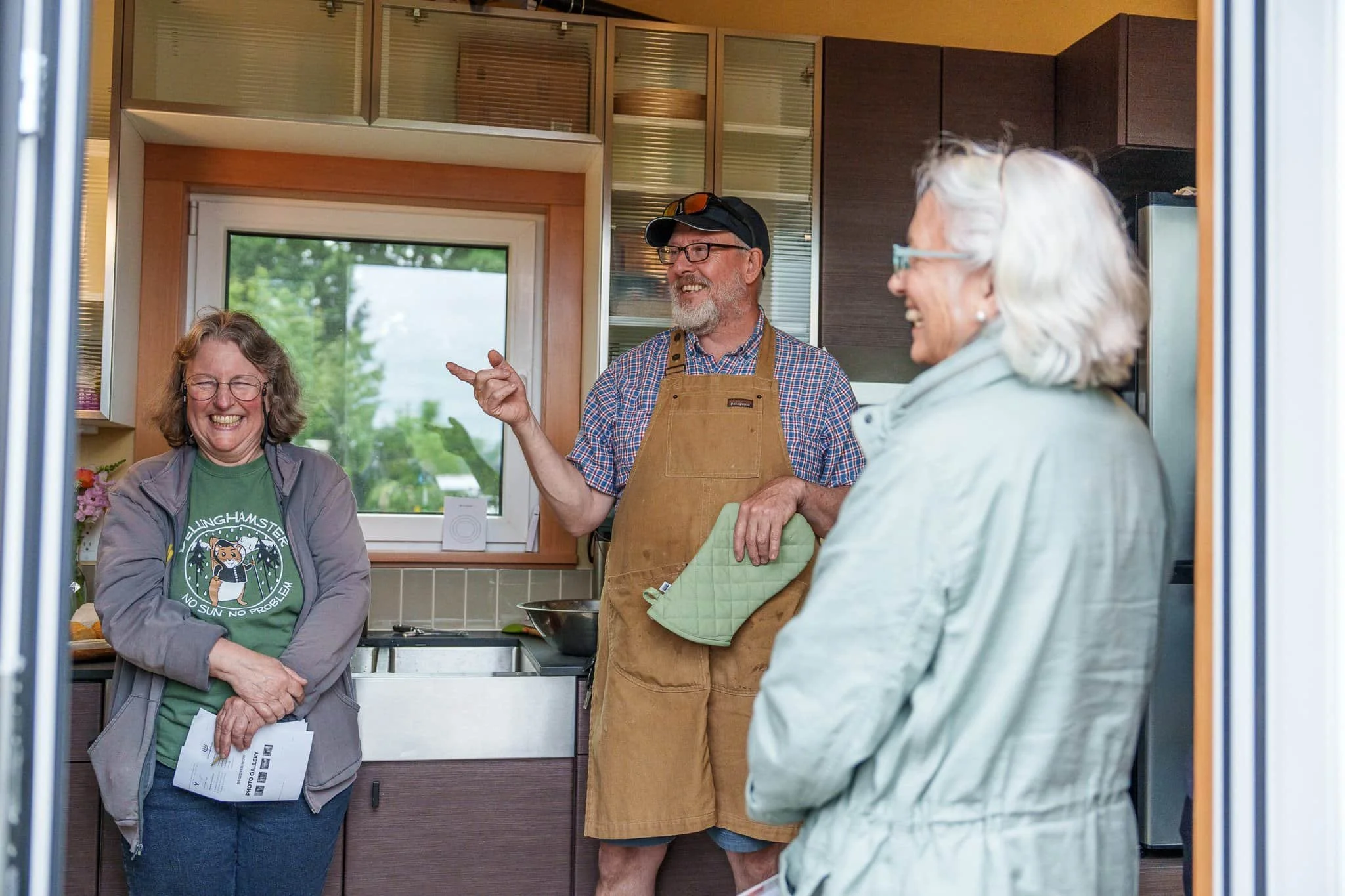 Three adults in a kitchen laugh together; one wears an apron and oven mitt, while two others stand nearby, smiling.
