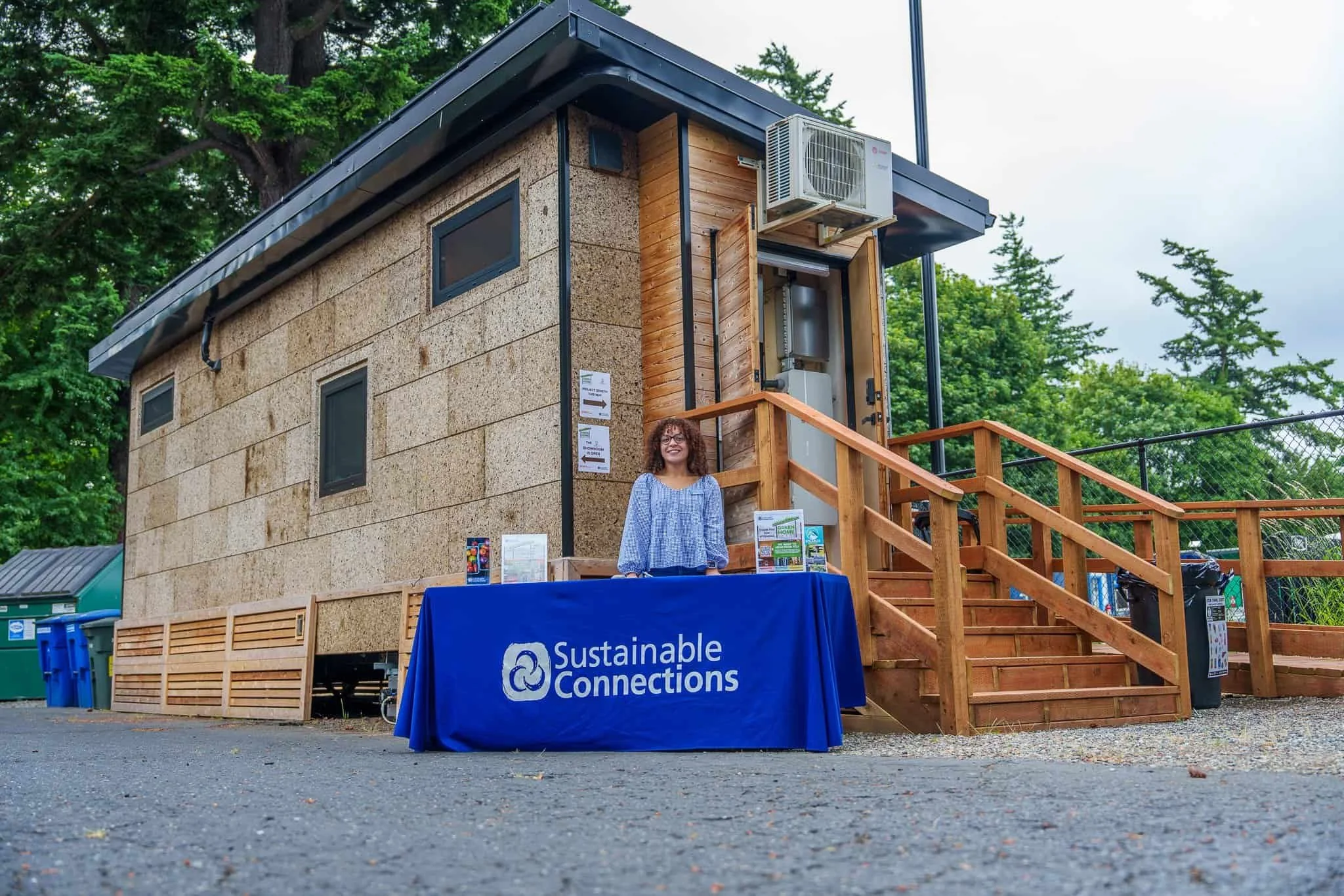 A woman stands behind a Sustainable Connections table in front of a small wooden building with trees in the background.