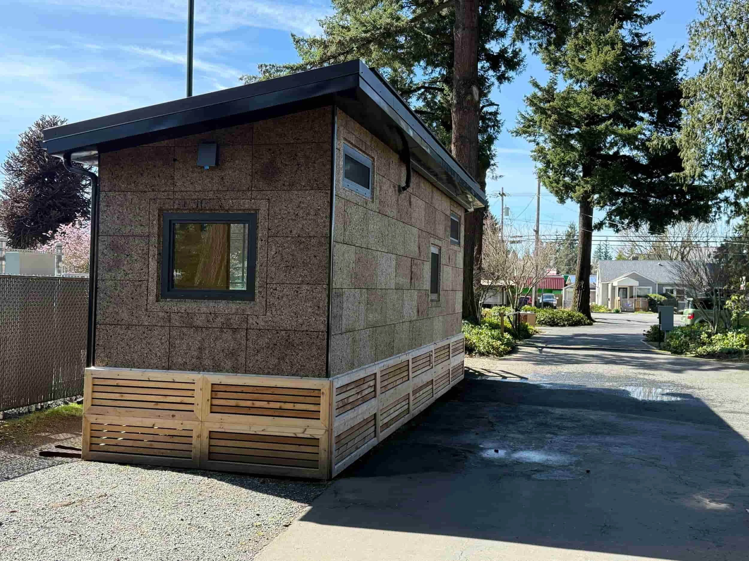 A small modern tiny house with wood siding and a single window, set on a sunny lot with trees and a fence.