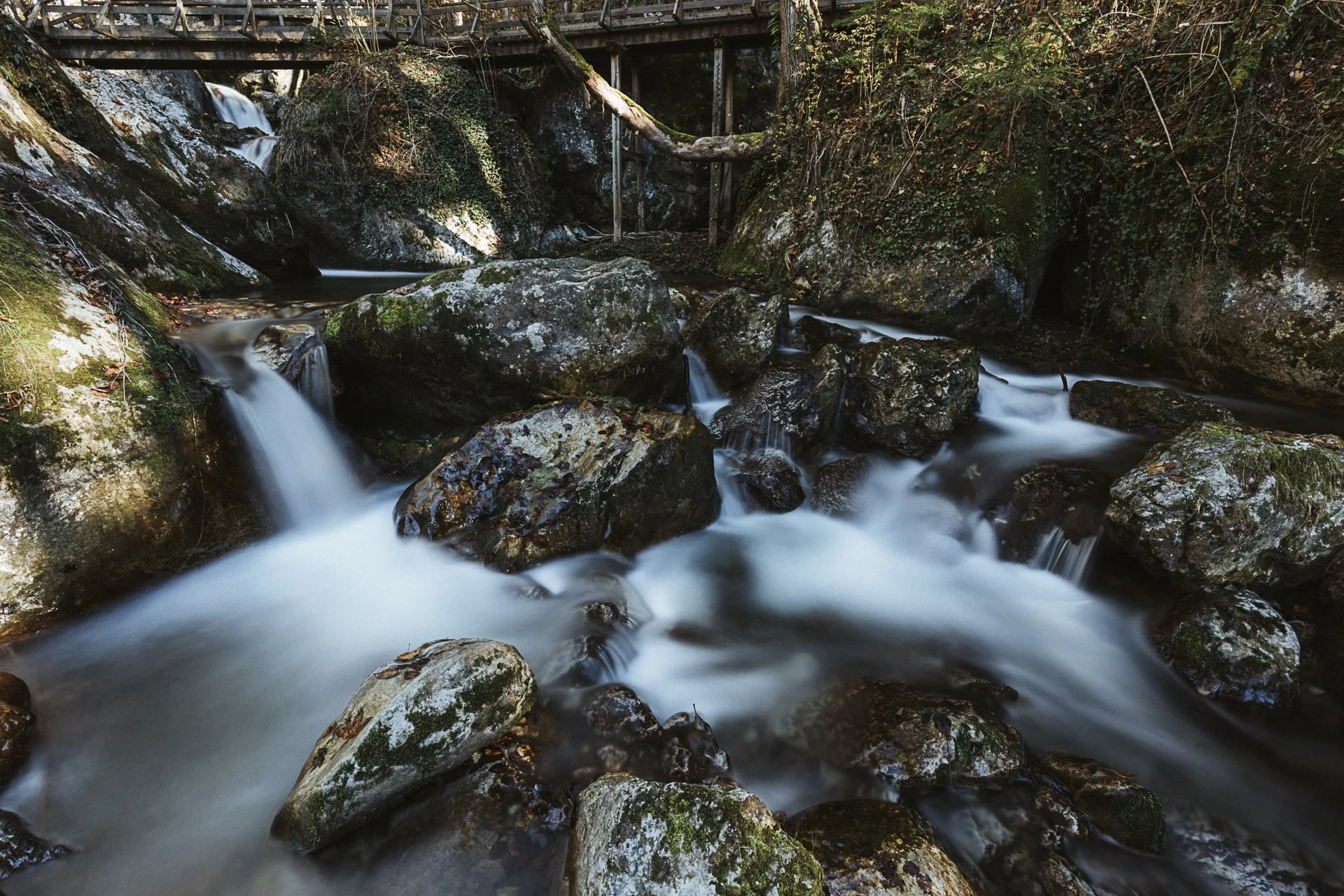Ein Bach fließt durch einen bewaldeten Bereich mit großen Steinen und Felsen, darüber eine Brücke aus Holz und Metall