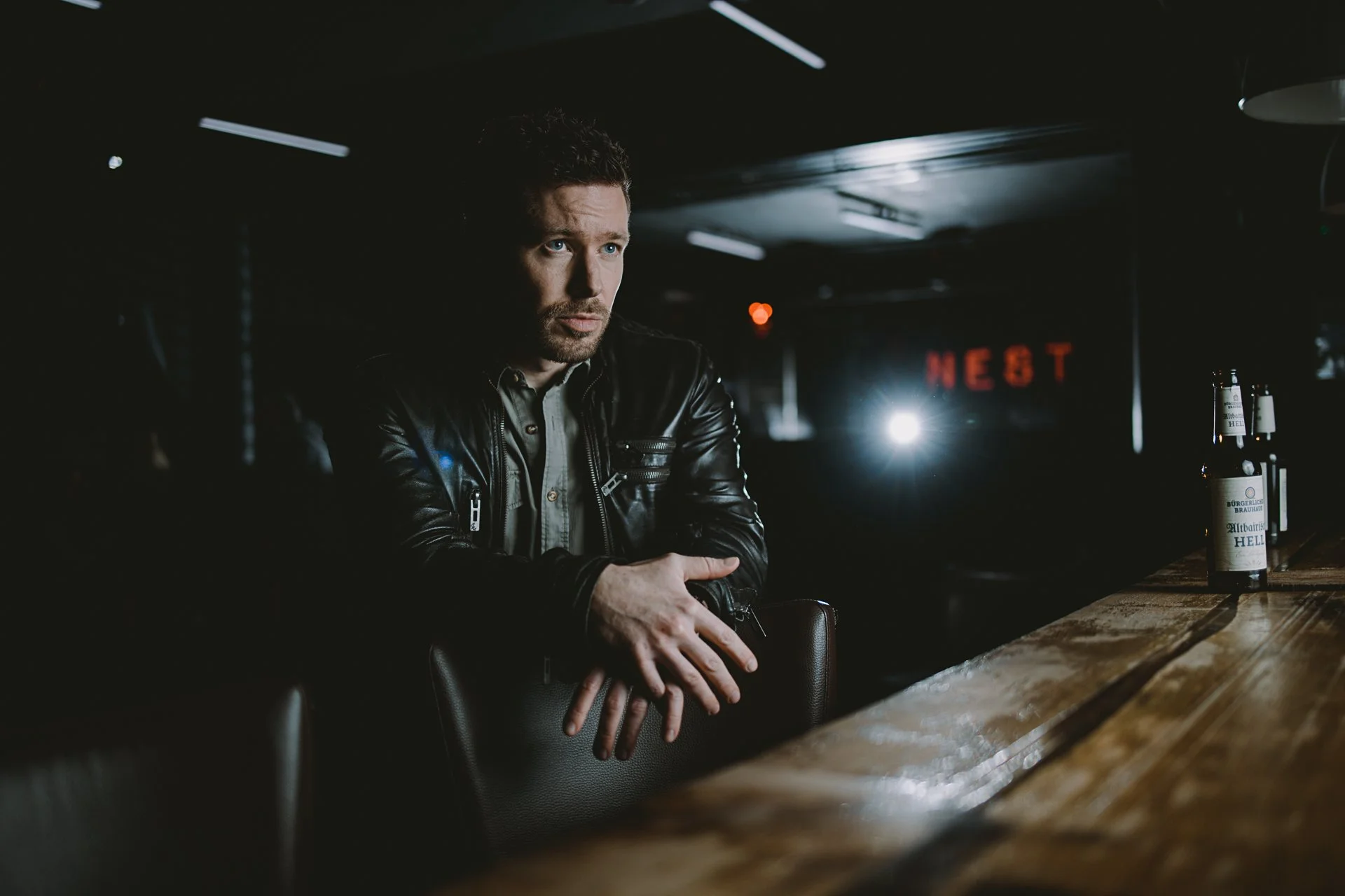 Ein Mann mit dunklem, lockigem Haar und einem Bart sitzt an einer Bar, trägt eine schwarze Lederjacke und schaut nachdenklich. Im Hintergrund sind leuchtende Neonbuchstaben, die teilweise sichtbar sind, sowie ein Lichtermeer in einem dunklen Raum. Au