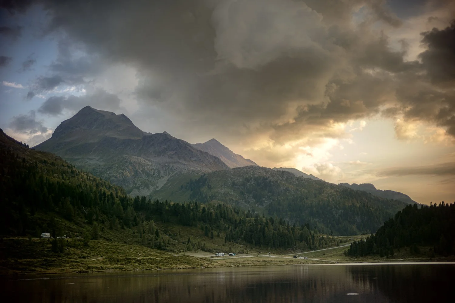 Ein Bergsee im Tal, umgeben von bewaldeten Hügeln und hohen Bergen, mit dunklen Wolken am Himmel und leichtem Licht am Horizont.