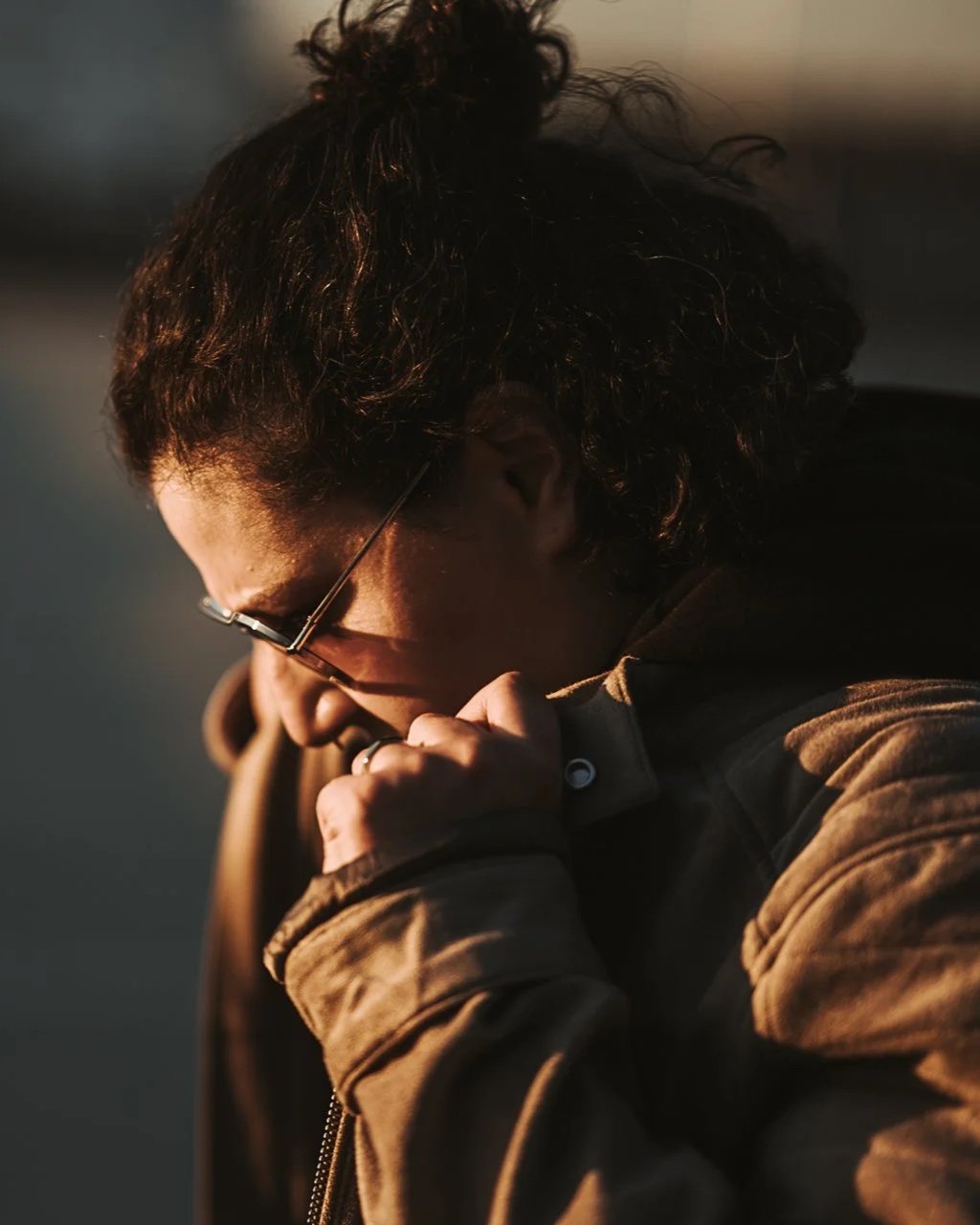 Junge Frau mit Brille und dunklen, lockigen Haaren sitzt nachdenklich und hält die Hand vor den Mund bei Sonnenuntergang.