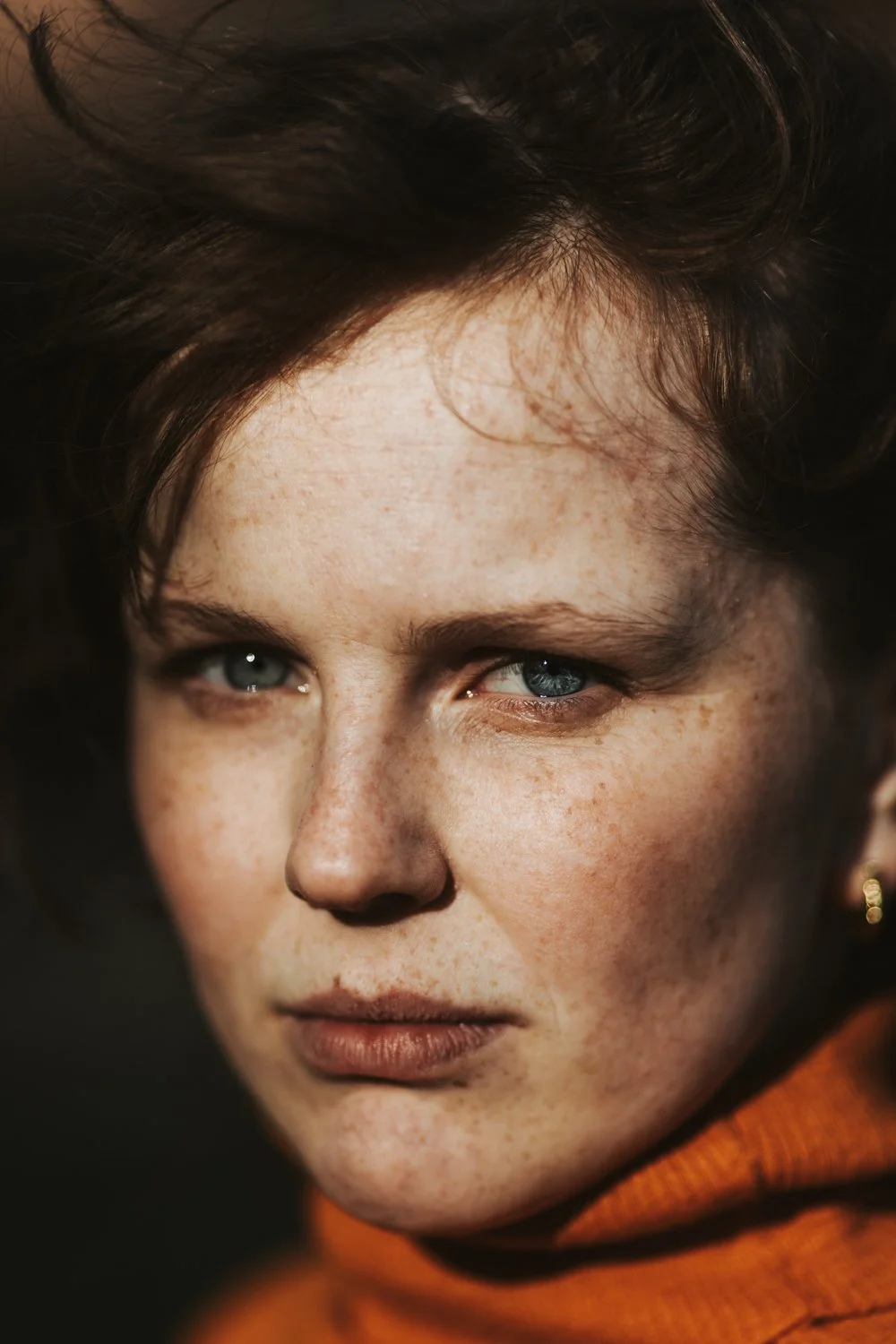 Nahaufnahme eines jungen Mädchens mit blauen Augen, braunen Locken, Sommersprossen und einem orangefarbenen Pullover.