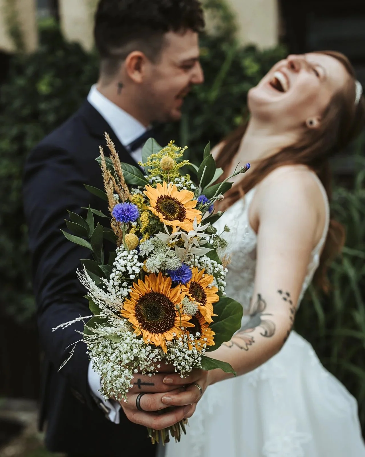 Ein junges Brautpaar bei der Hochzeit, die Braut hält einen großen Blumenstrauß mit Sonnenblumen, Kornblumen und anderen Blumen. Beide lachen freudig, der Bräutigam trägt einen schwarzen Anzug und die Braut ein weißes Kleid. Im Hintergrund ist Vegetation sichtbar.