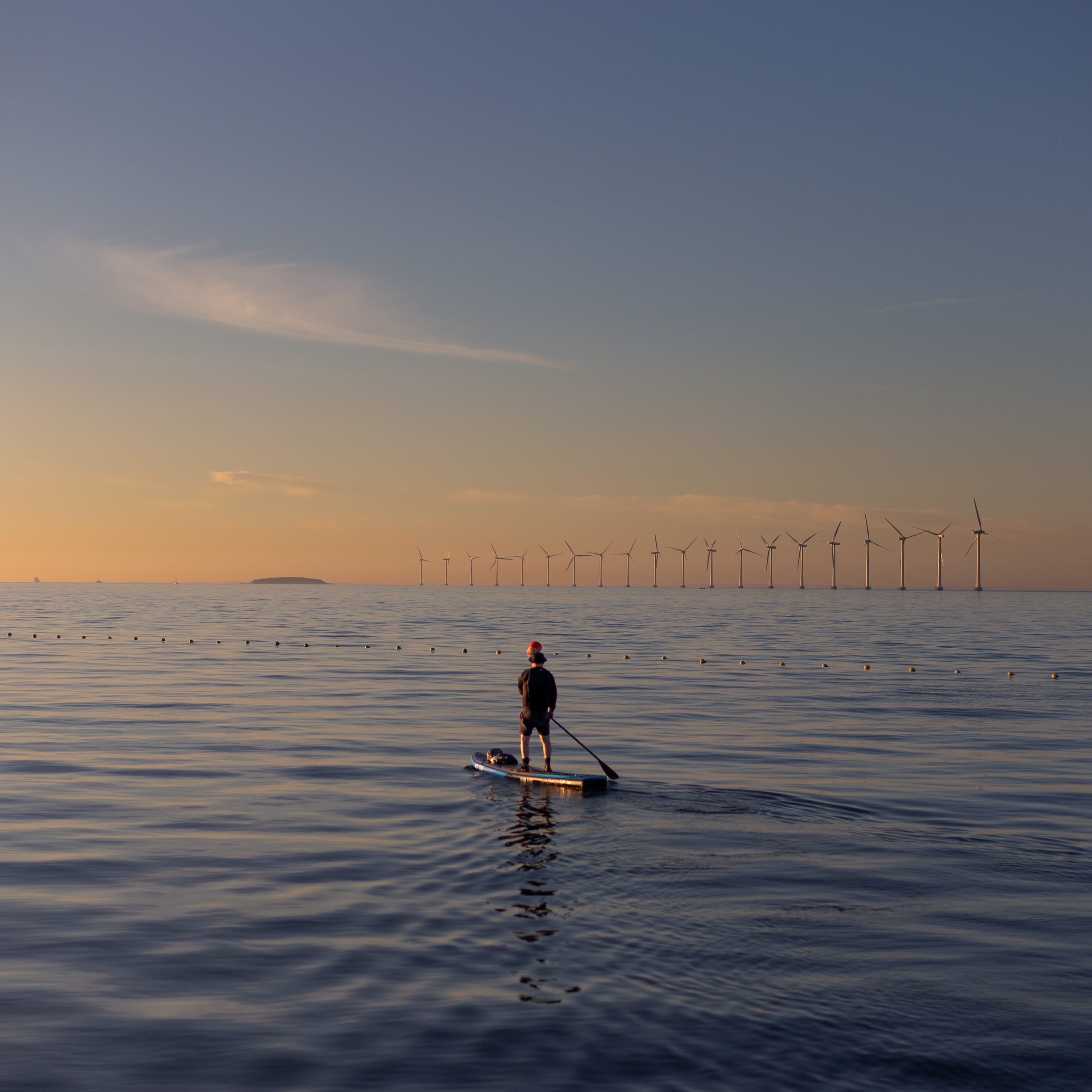 Ein Mensch steht auf einem Paddle-Board im Wasser bei Sonnenuntergang, mit Windkraftanlagen im Hintergrund.