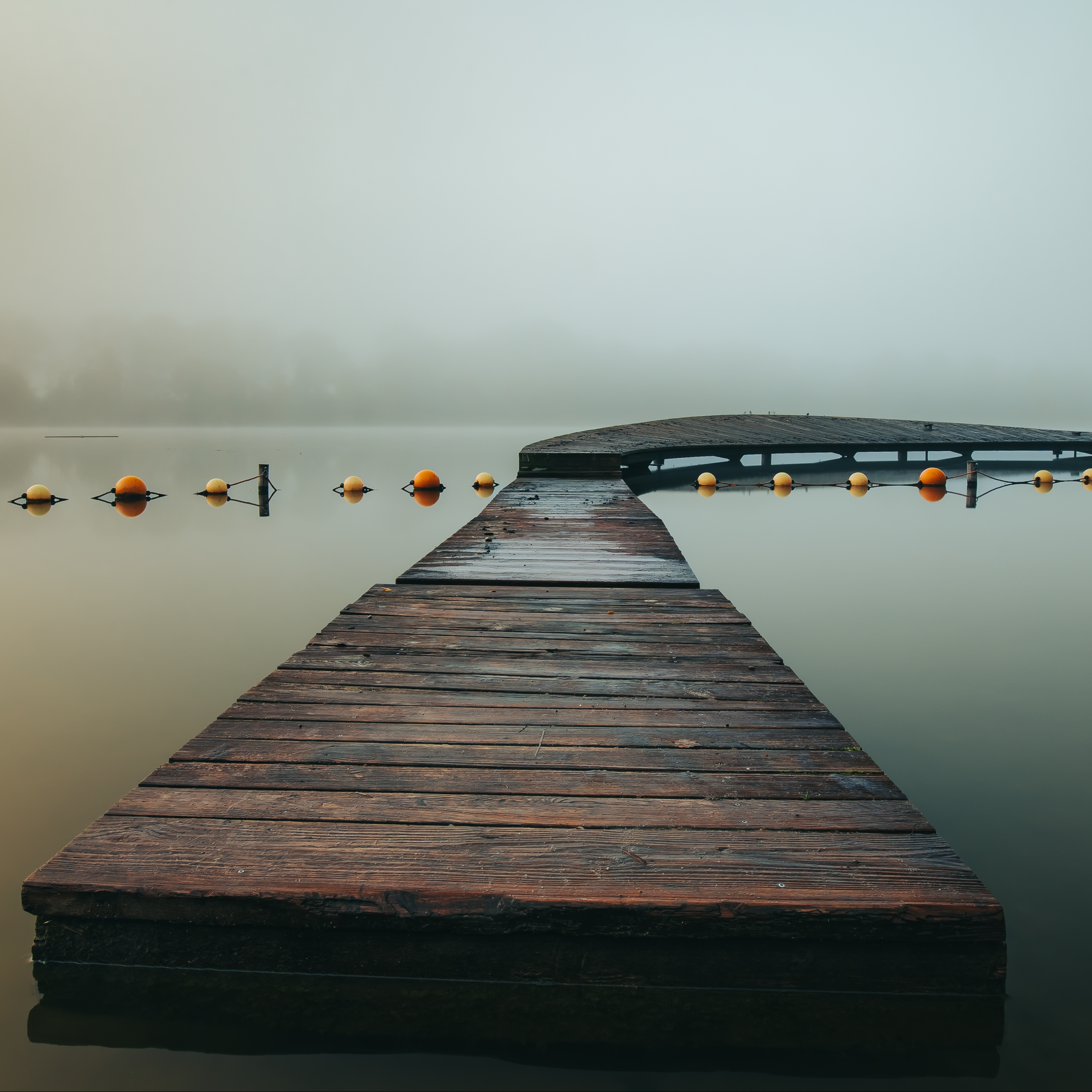 Ein Holzsteg, der ins Wasser führt, mit Bojen an der Seite, an einem bewölkten Tag. Im Hintergrund ist Wasser und Himmel sichtbar.
