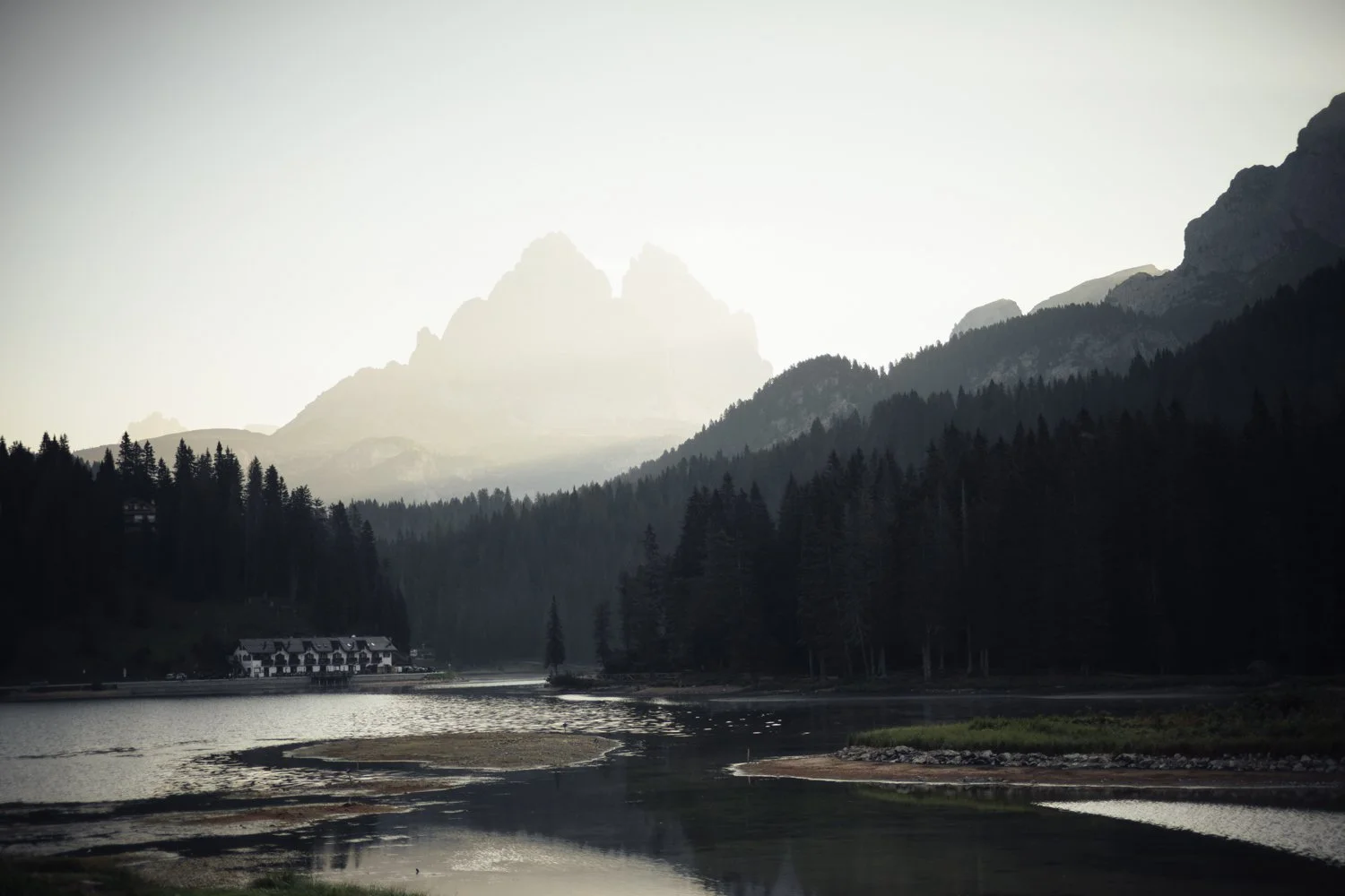 Berglandschaft mit Wald, Fluss und Häuser im Vordergrund, im Hintergrund Berge unter bewölktem Himmel.