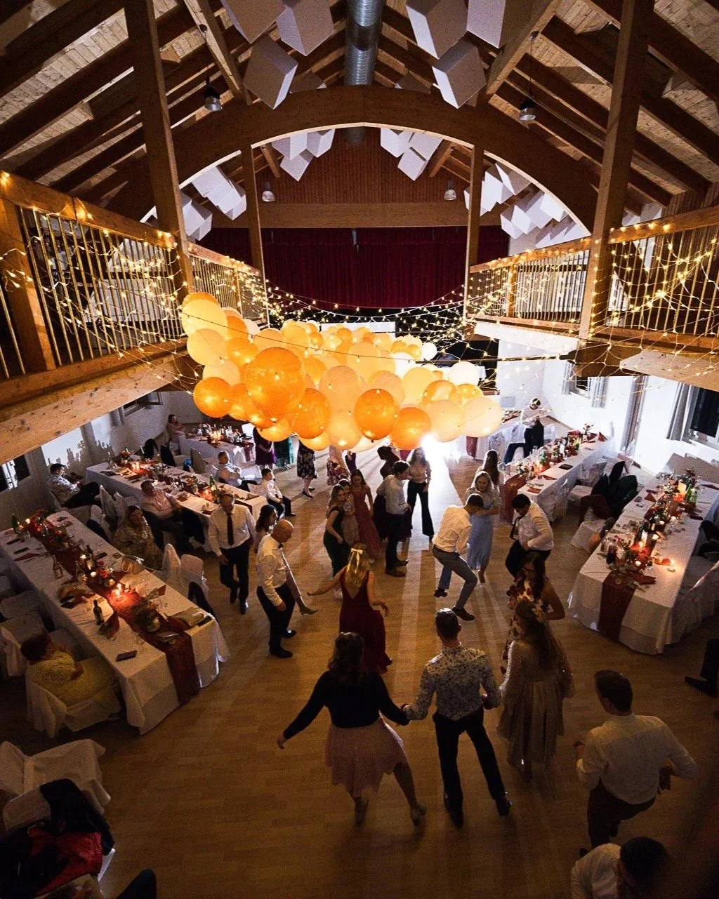 Eine Festhalle mit einem Tanzboden, gedeckten Tischen und einer Dekoration aus Lichterketten und Luftballons, auf der Tanzfläche tanzen Menschen.