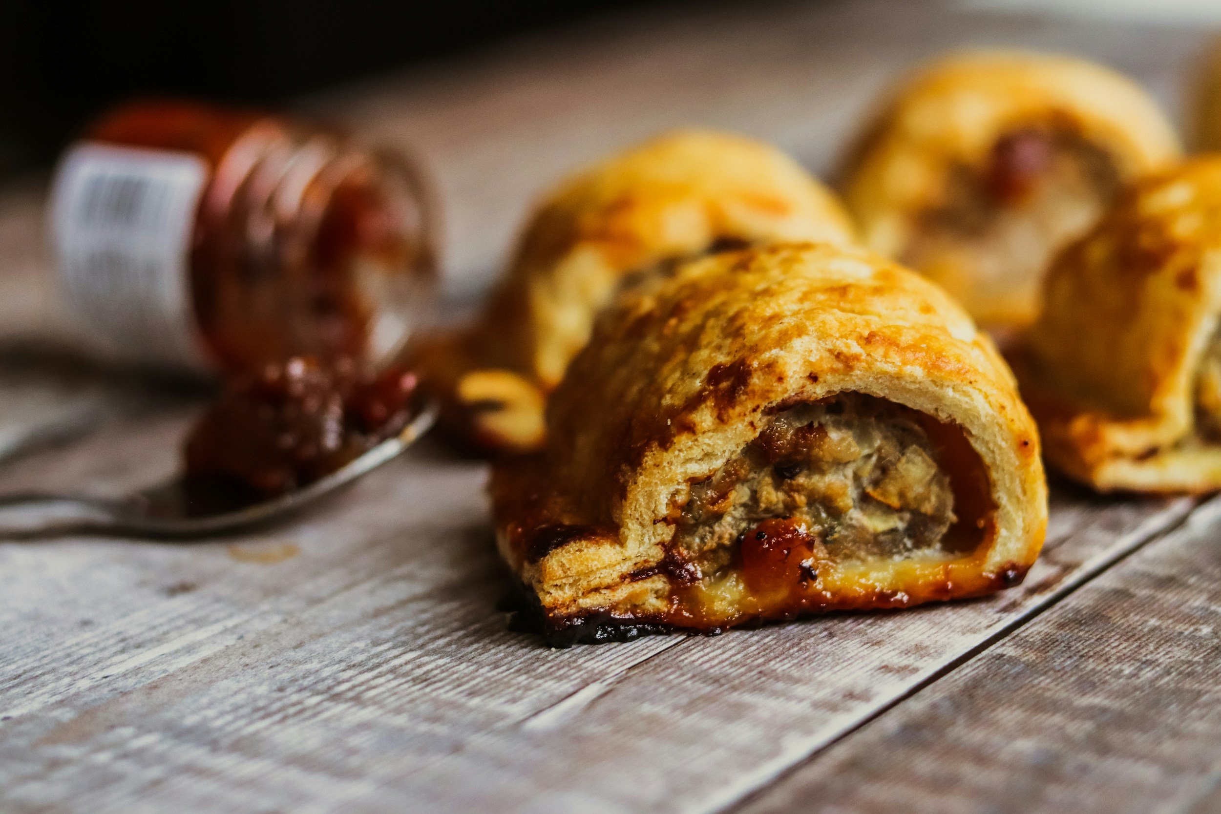 A baked pastry with a meat and vegetable filling, partially sliced open on a rustic wooden surface, with a jar of jam and a spoon nearby.