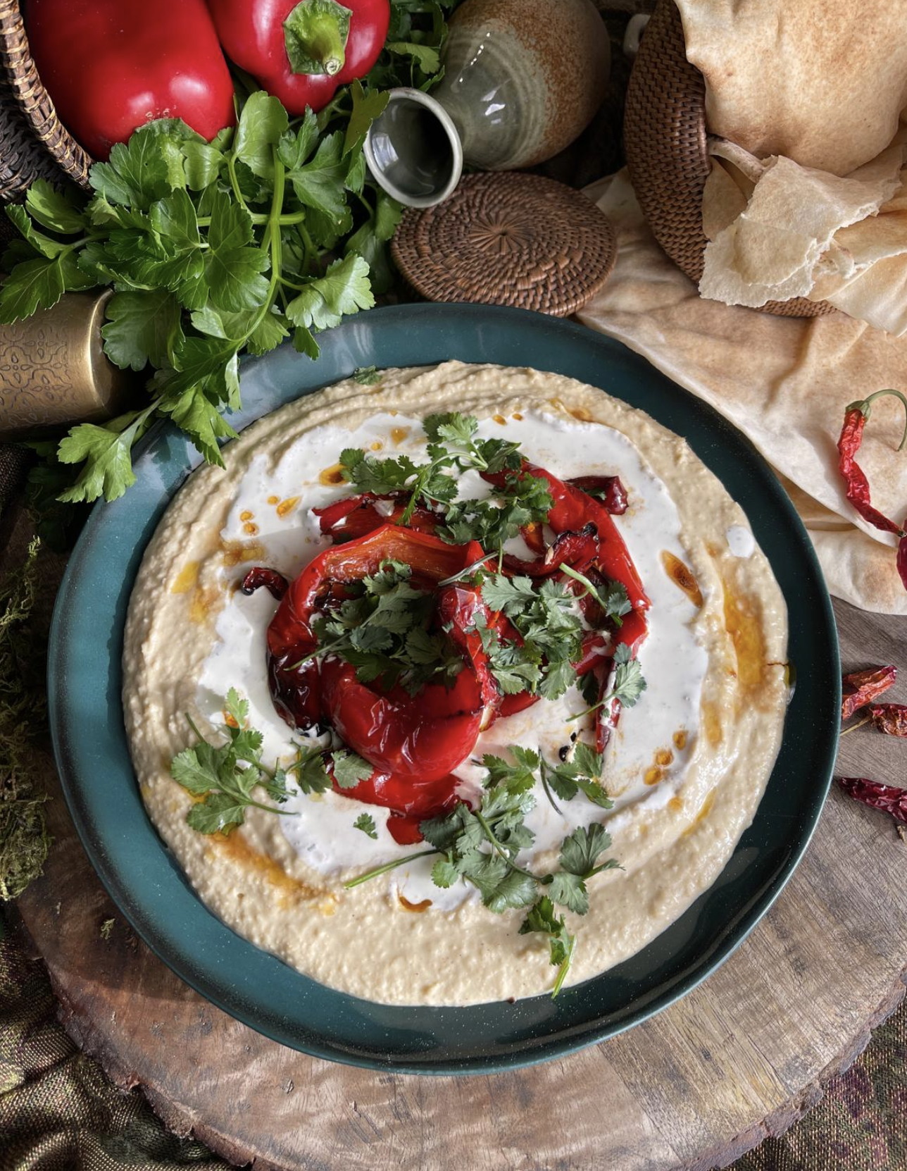 Plate of creamy hummus topped with roasted red peppers, fresh cilantro, and drizzled with olive oil, surrounded by fresh herbs, peppers, and bread.