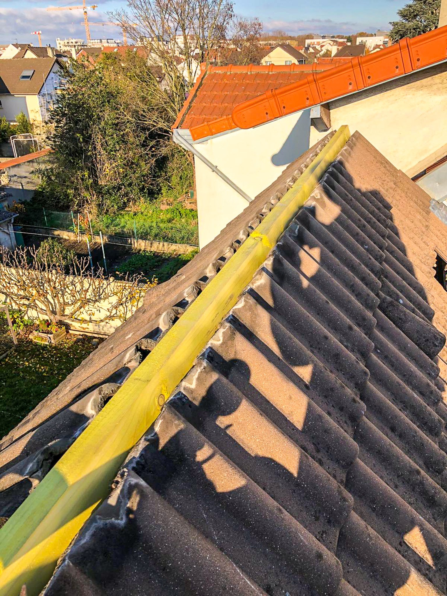 Vue depuis un toit montrant une ligne de support jaune pour la réalisation de travaux de couverture, avec des tuiles noires, une maison avec un toit en tuiles orange et un paysage urbain en arrière-plan.