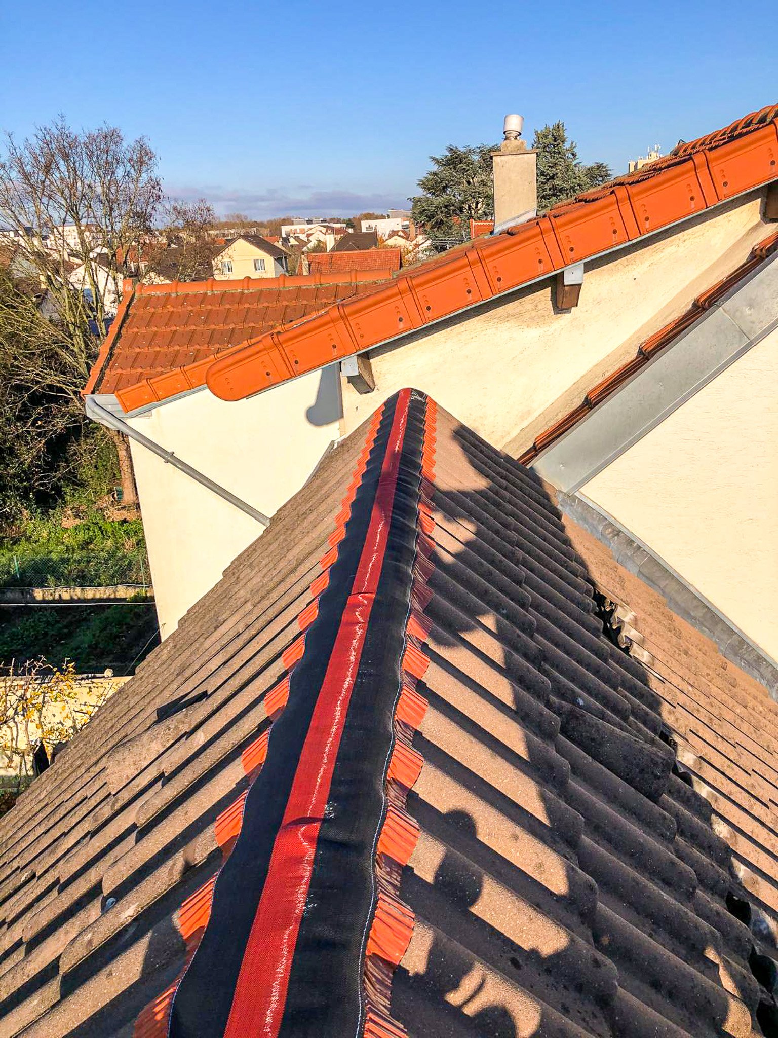 Vue sur plusieurs toitures de maisons avec des tuiles en terre cuite, dont certaines neuves de couleur orange et d'autres anciennes plus sombres, sous un ciel bleu clair.