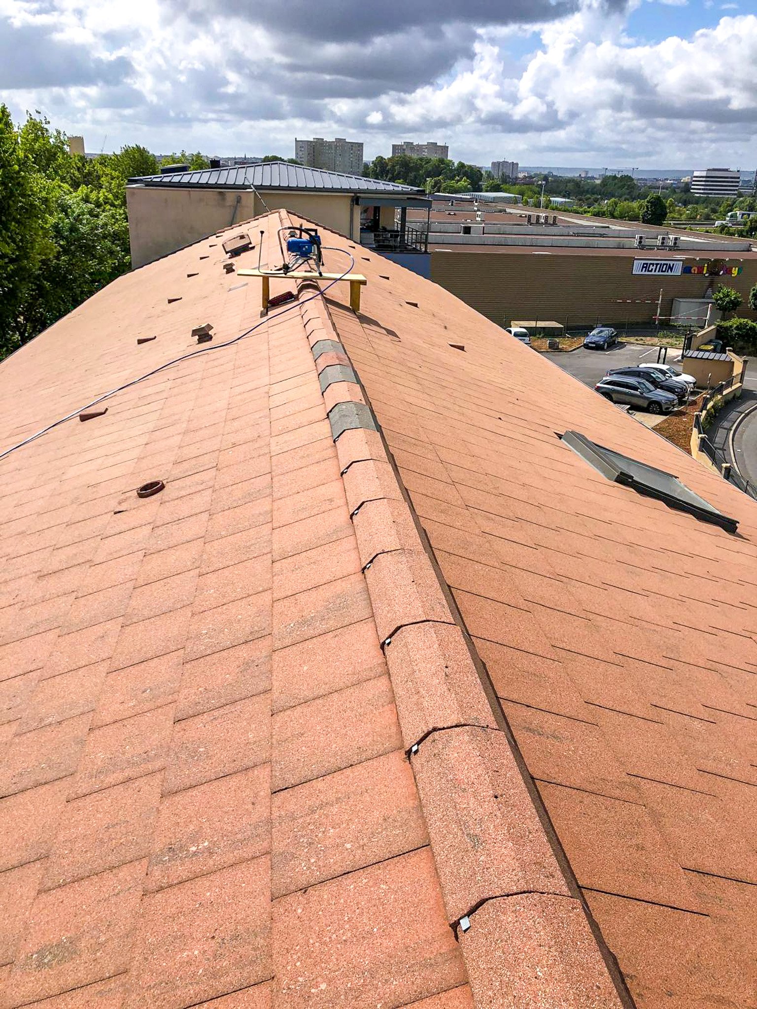 Vue du sommet d'une toiture en tuiles rouges, avec un équipement de travail posé dessus, dans un environnement urbain.