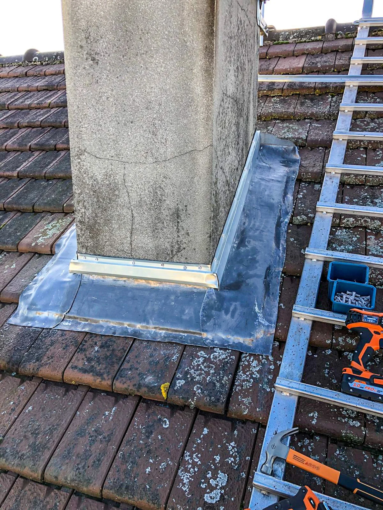 Travaux de rénovation sur une cheminée avec une nouvelle couverture en aluminium, outils à côté sur le toit en tuiles.