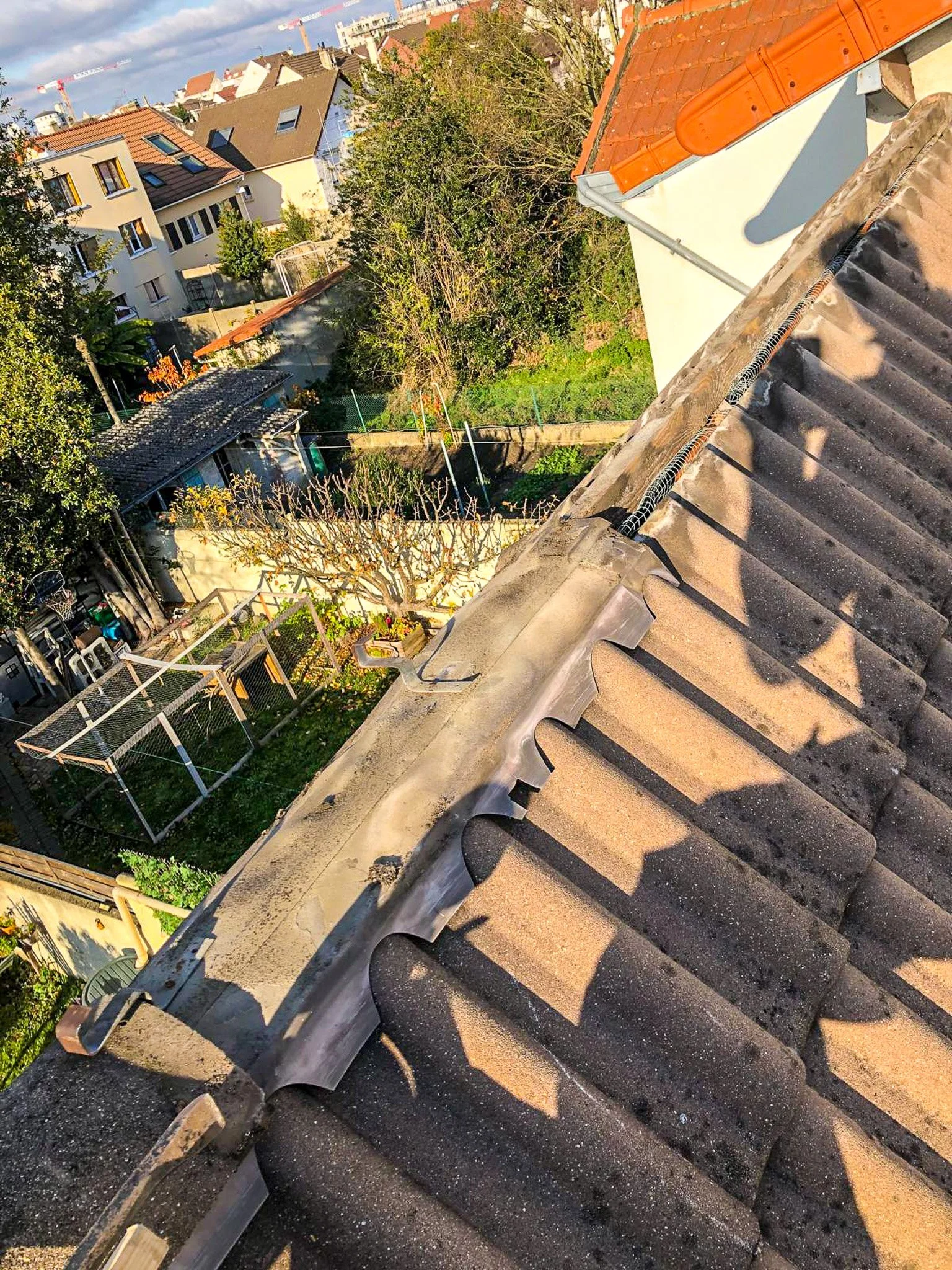 Vue aérienne d'un toit en tuiles avec travaux de réparation en cours, des maisons, des arbres et un jardin visibles en arrière-plan.