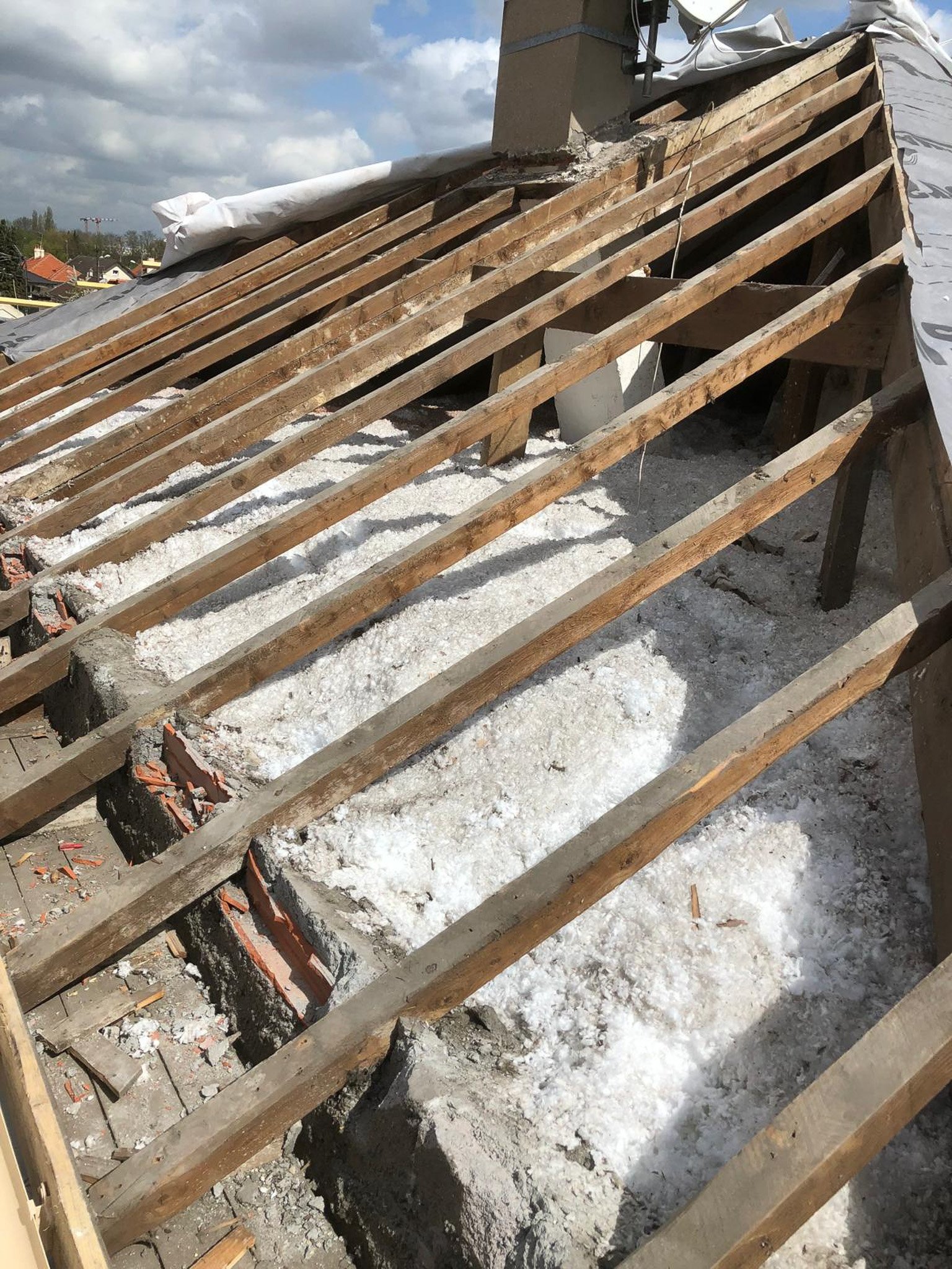Structure de toiture en construction avec poutres en bois, isolant blanc, et cheminée en brique, sous un ciel partiellement nuageux.