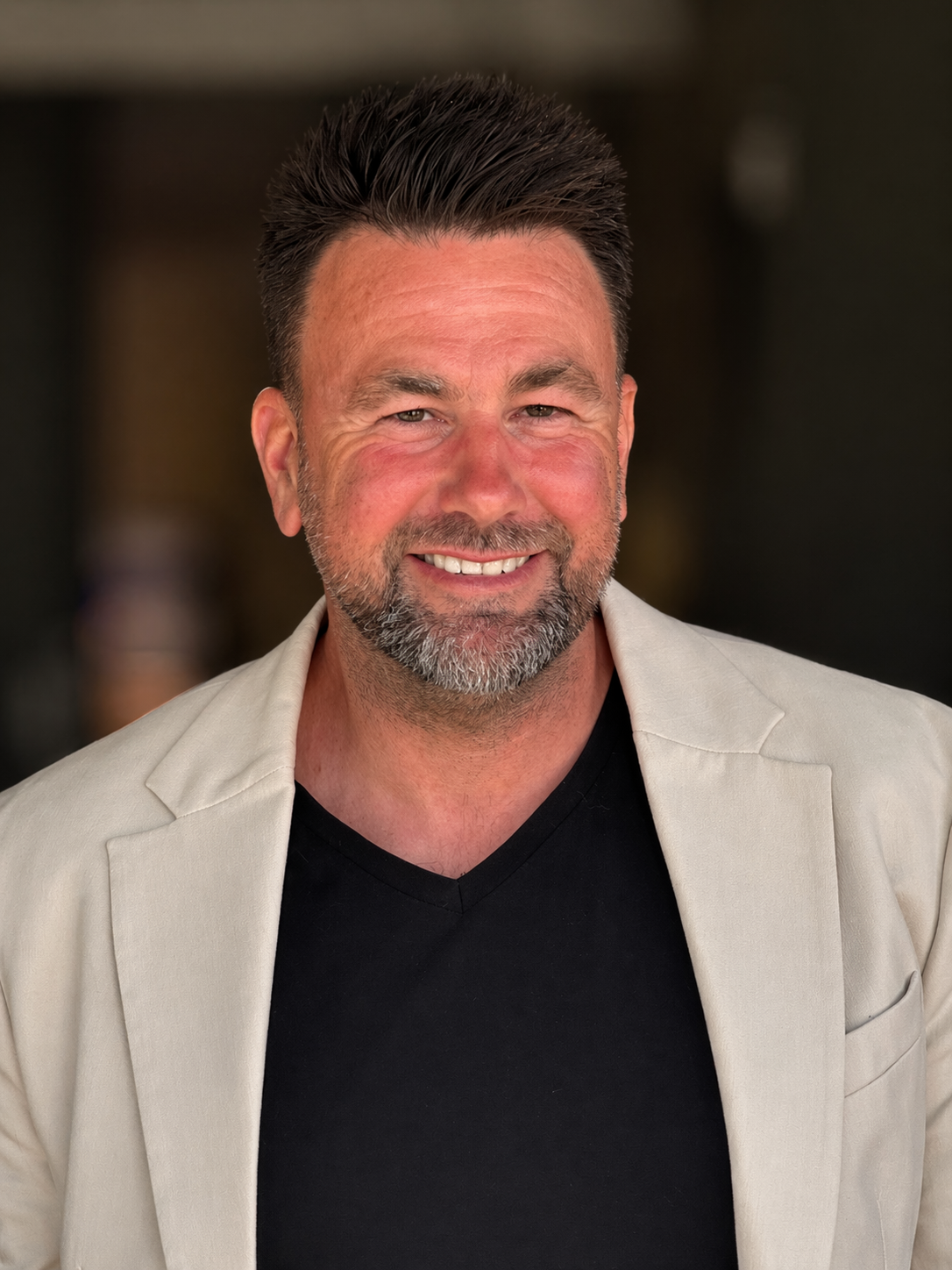 A smiling middle-aged man with short dark hair, a beard, and a mustache, wearing a black V-neck shirt and a beige blazer, standing outdoors with a blurred background.