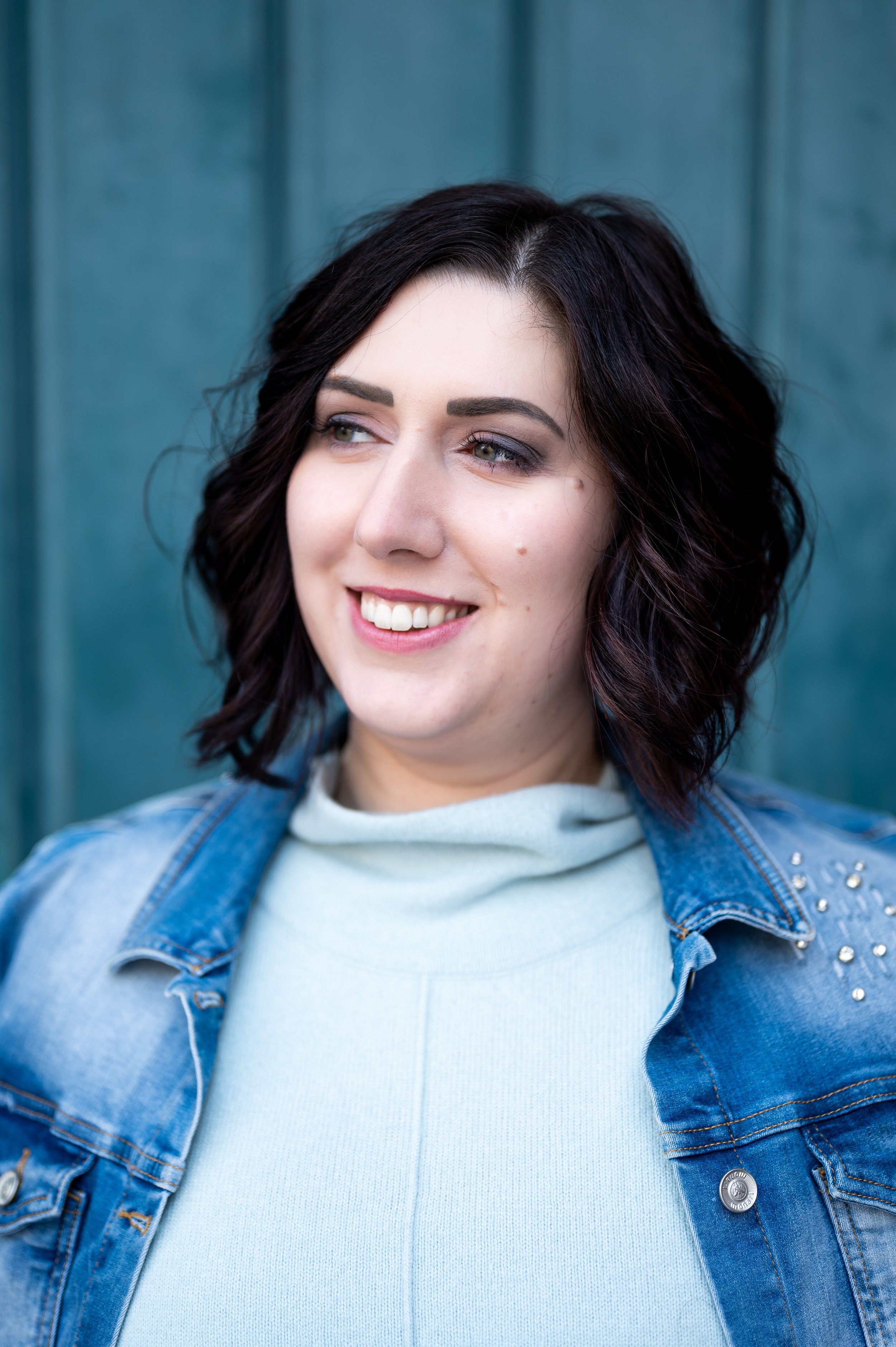 Junge Frau mit dunklen, gewellten Haaren, lächelnd, trägt ein hellblaues T-Shirt und eine Denim-Jacke. vor einer blauen Holzwand.