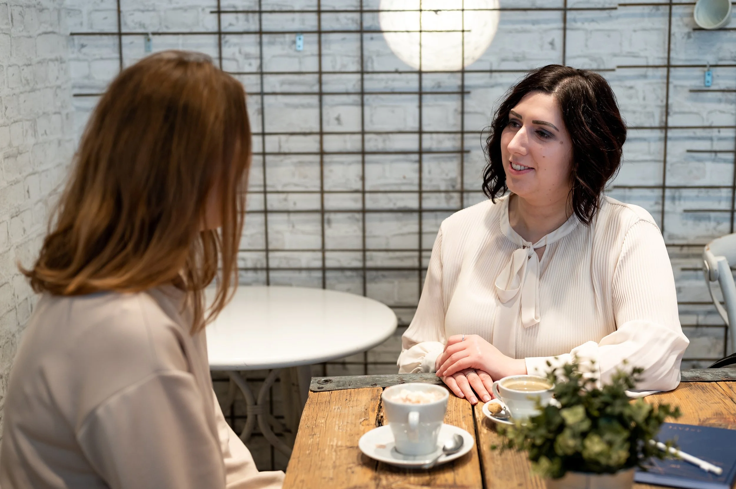 Zwei Frauen führen ein Gespräch in einem Café, beide sitzen an einem Holztisch mit Kaffee und einem Blumenstrauß.