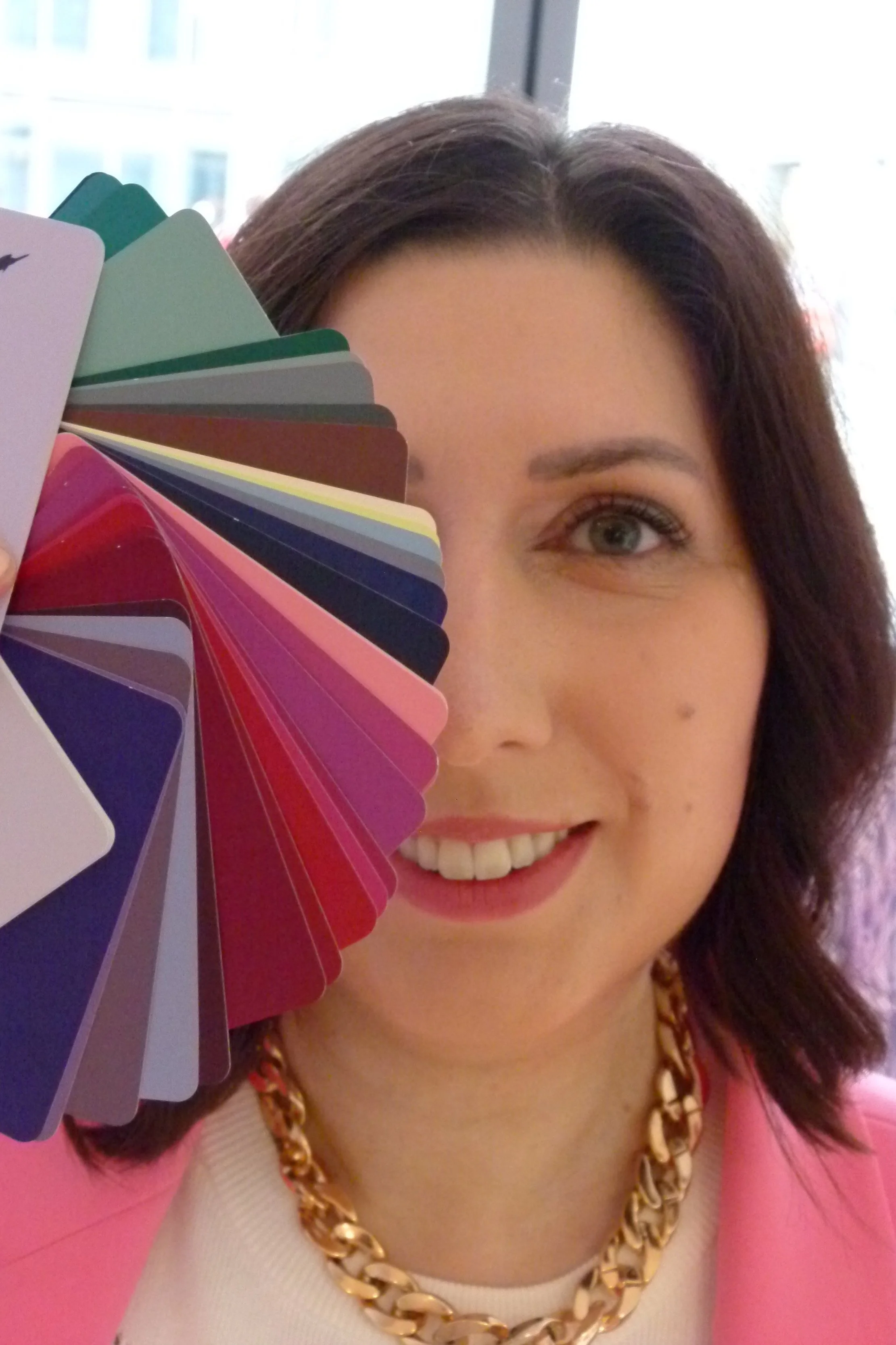 Frau mit braunen Haaren, blauer Augen, lachend, trägt pinke Jacke und goldene Kette, hält Farbkarte vor das Gesicht.